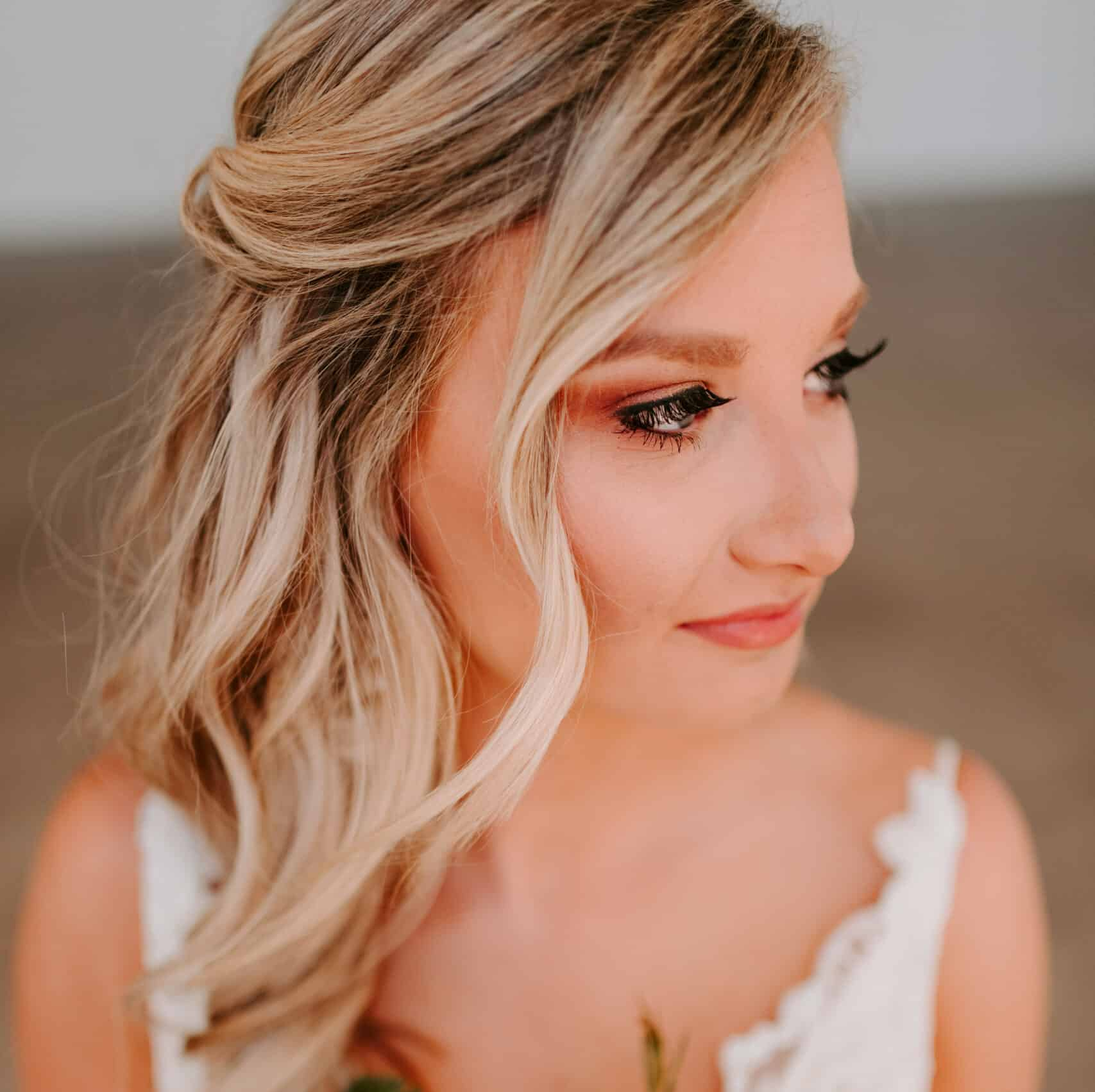 Blonde woman with wavy hair, half-updo, wearing a white dress. Looking right, soft makeup.
