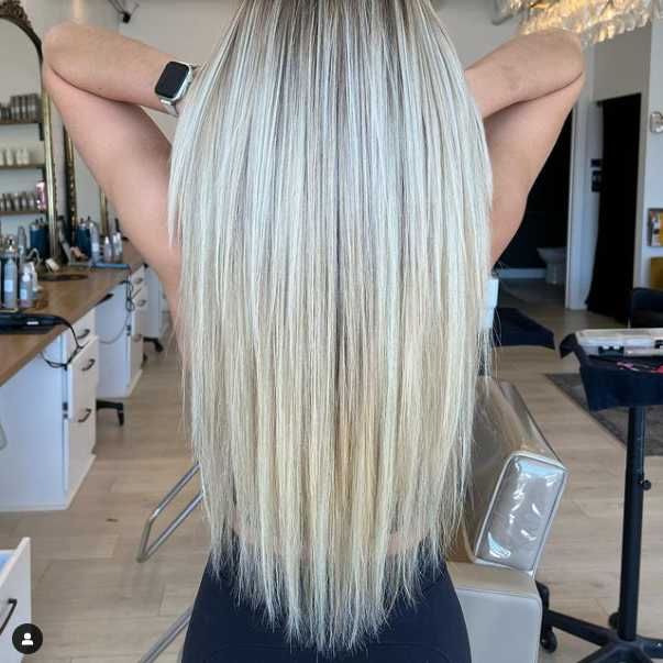 Woman with long, straight blonde hair, arms raised behind her head, at a salon.