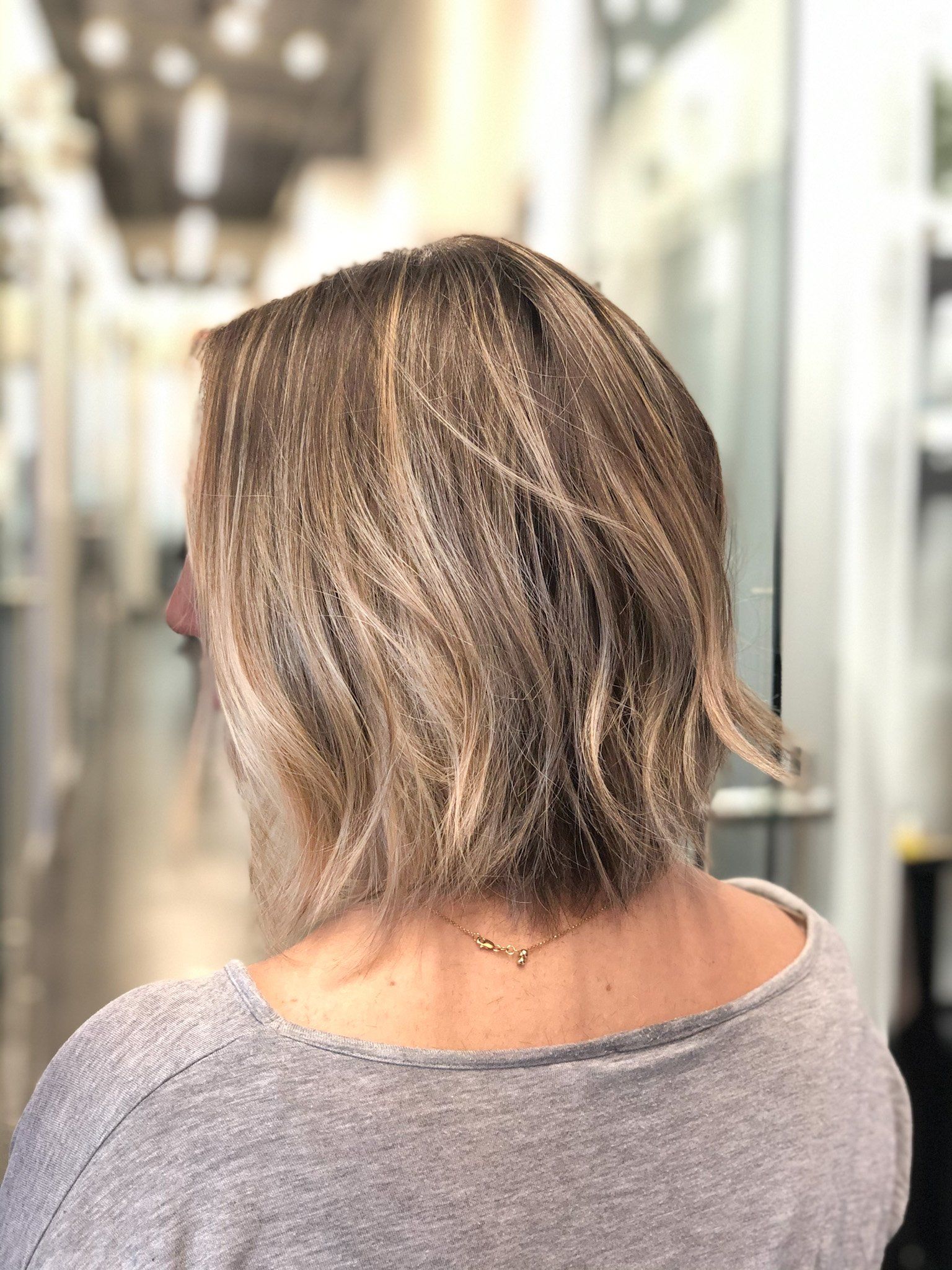 Blonde hair bob, wavy texture, back view. Wearing a gray sweater, standing indoors near glass doors.