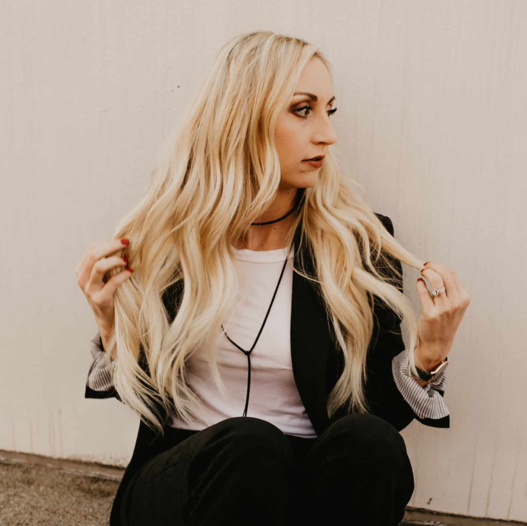 Woman with long blonde hair, in a black blazer and white shirt, looking to the side, touching her hair, sitting.