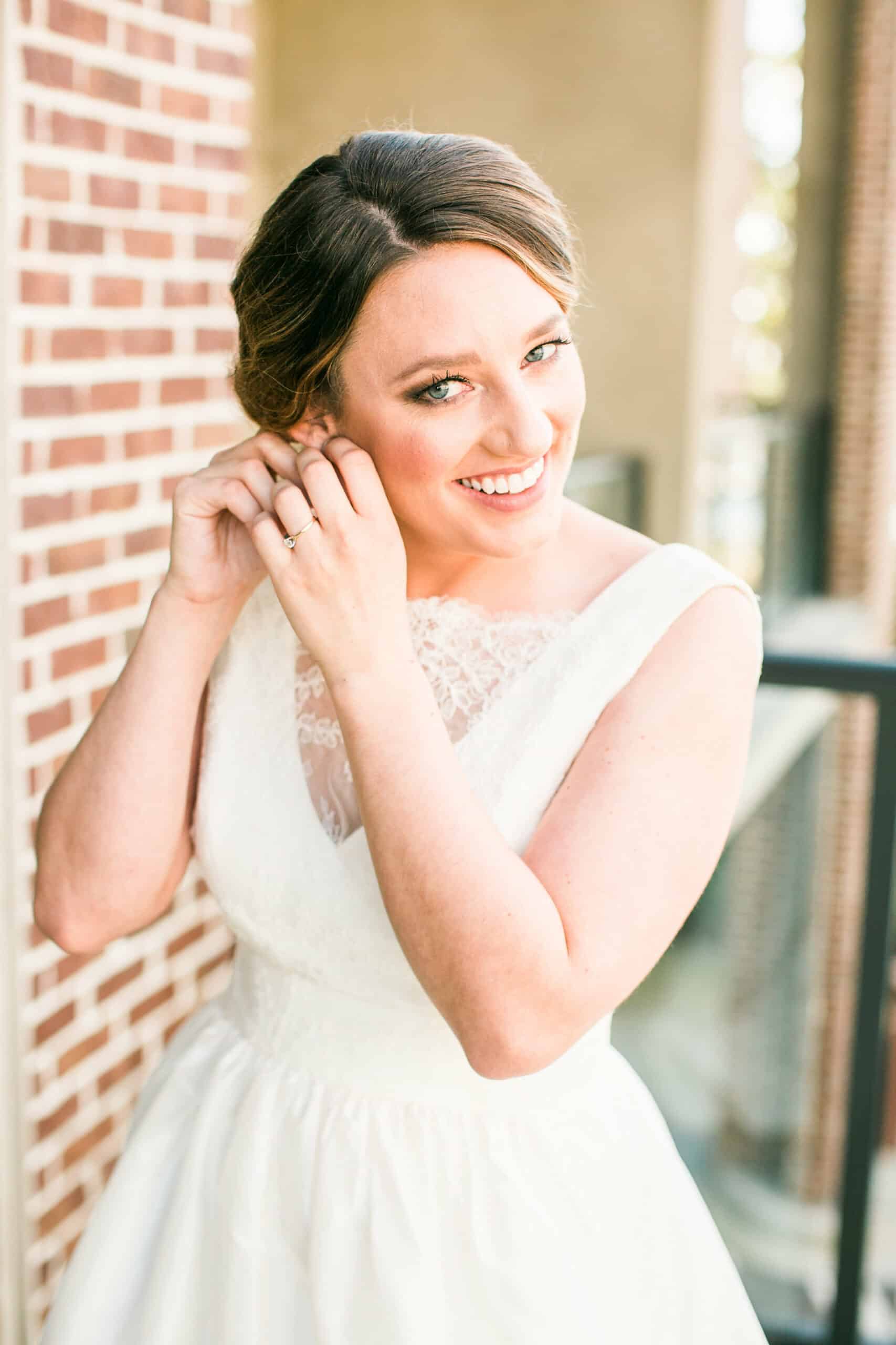 Bride in a white dress puts on earring, smiling near a brick wall.