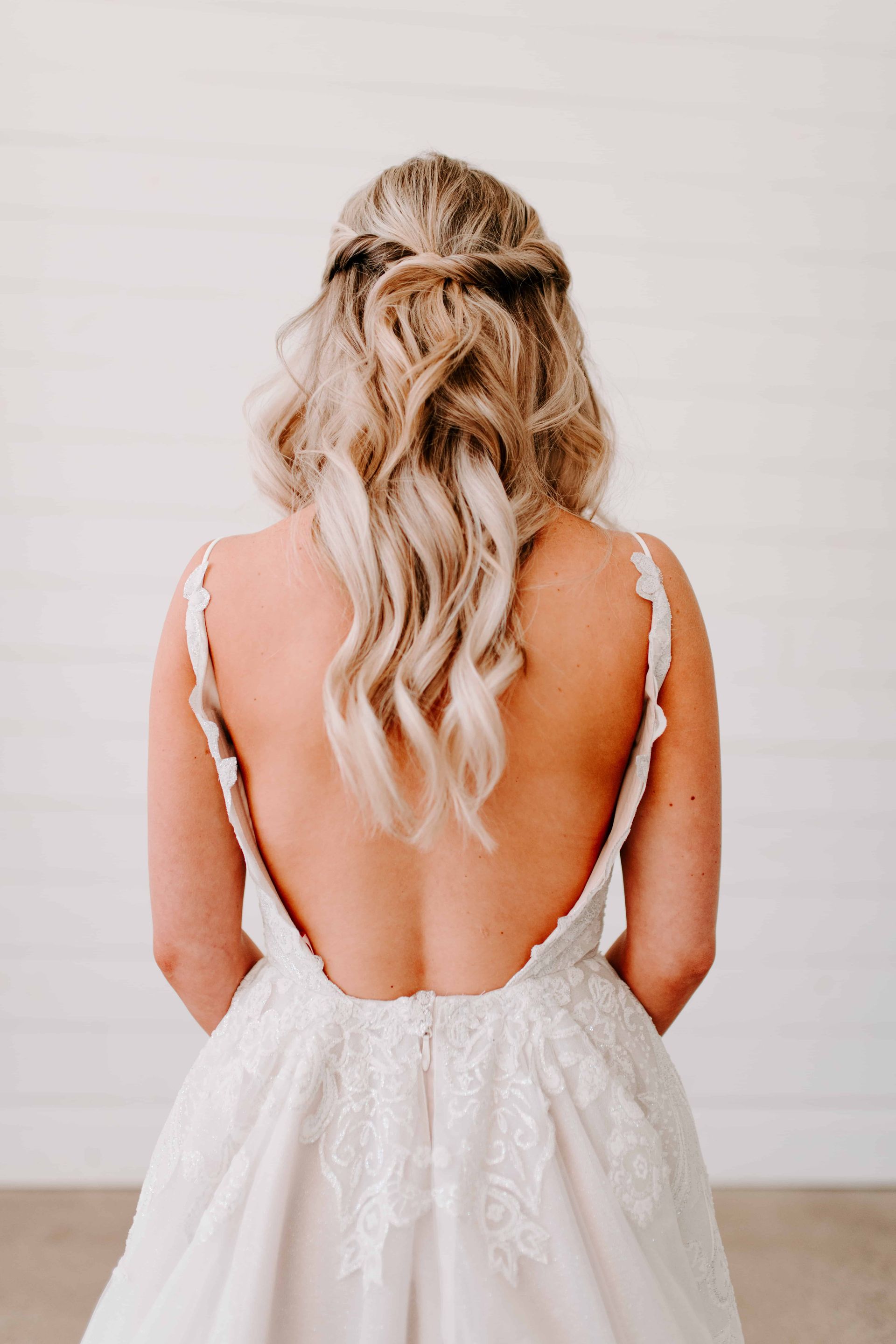 Woman in a wedding dress with a low back, looking away; blonde, wavy hair pulled half up.