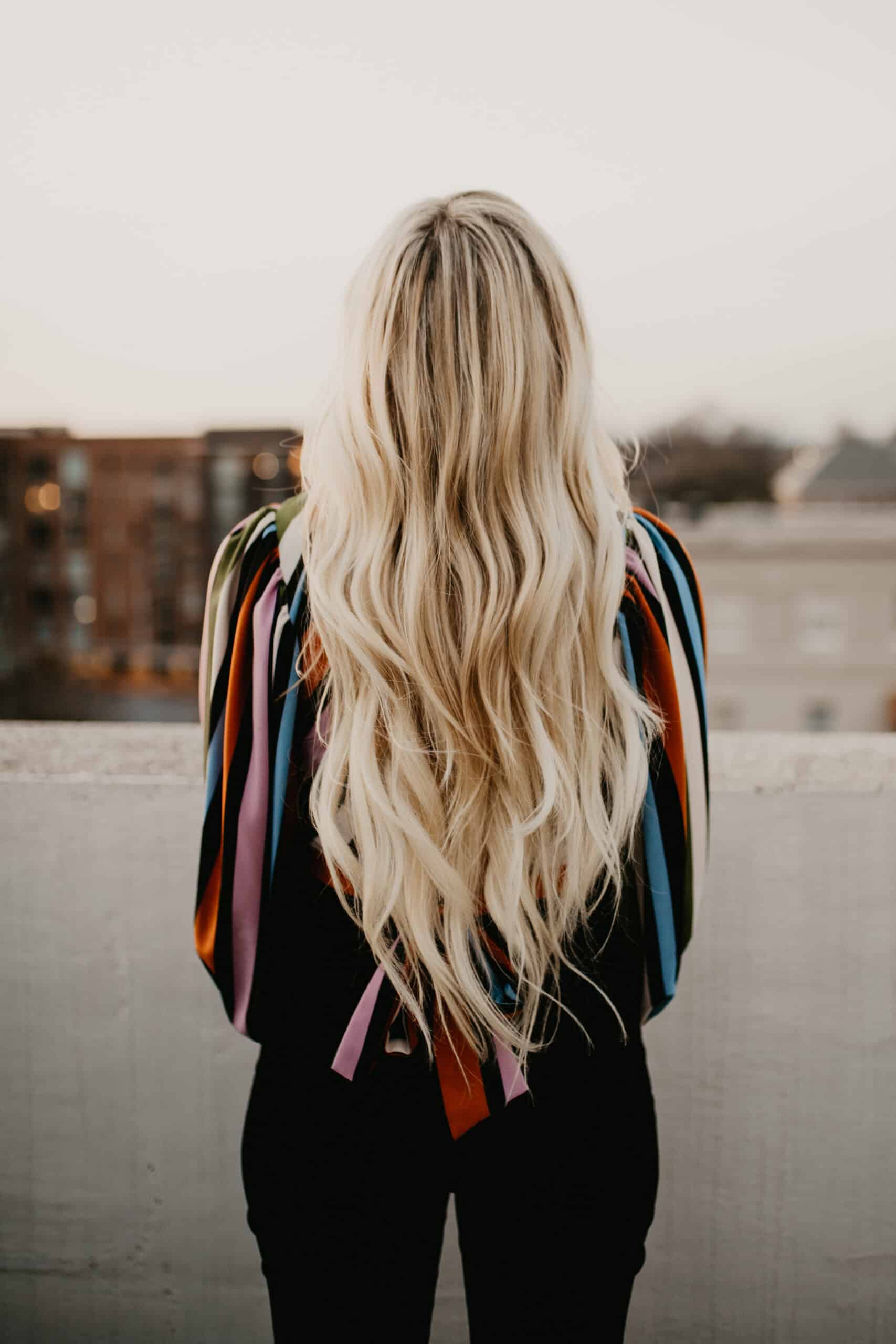 Woman with long blonde hair, standing with back to the camera. Striped top, black pants, on a rooftop overlooking city.