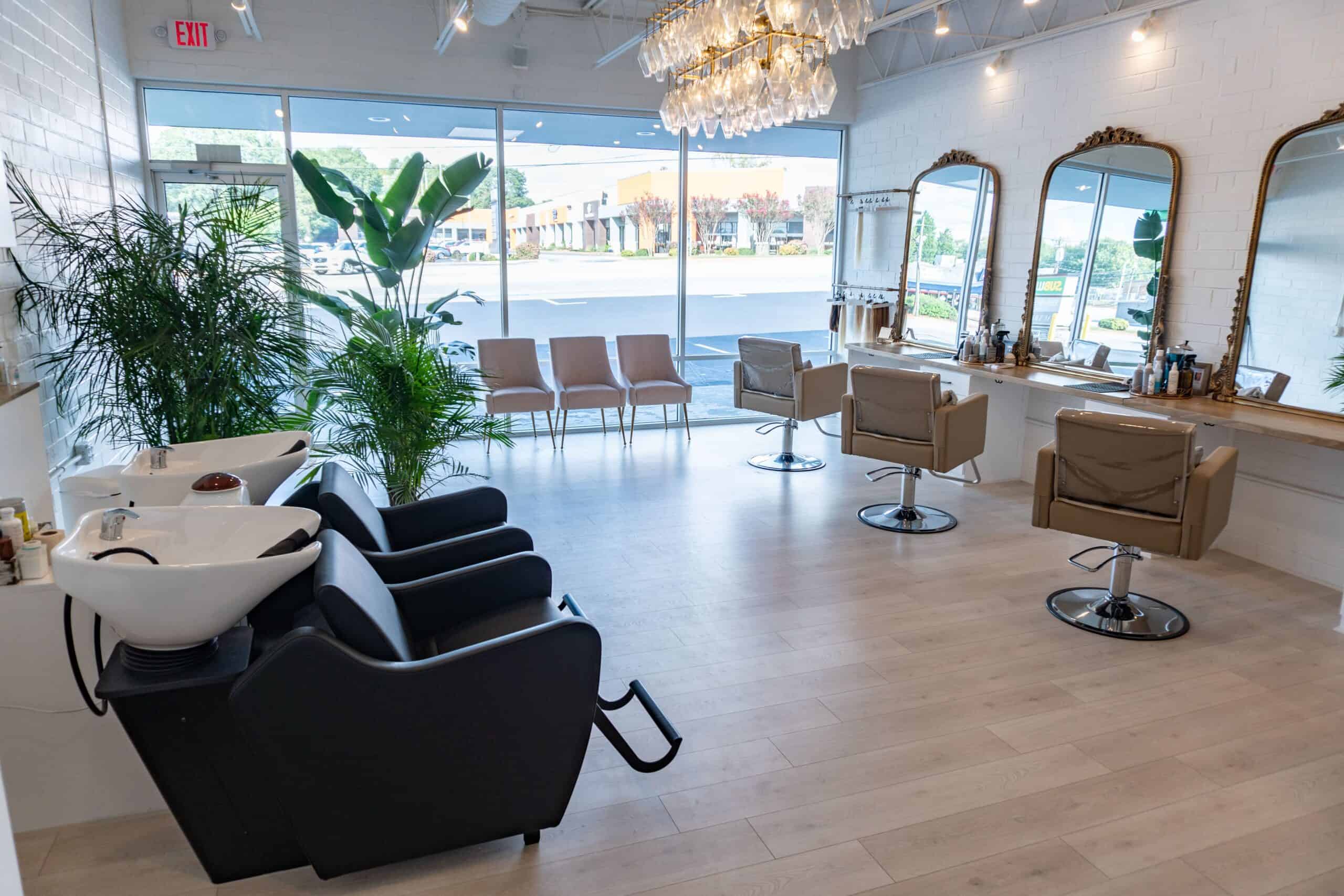 Hair salon interior with styling stations, wash basin, chairs, large windows, and a chandelier.