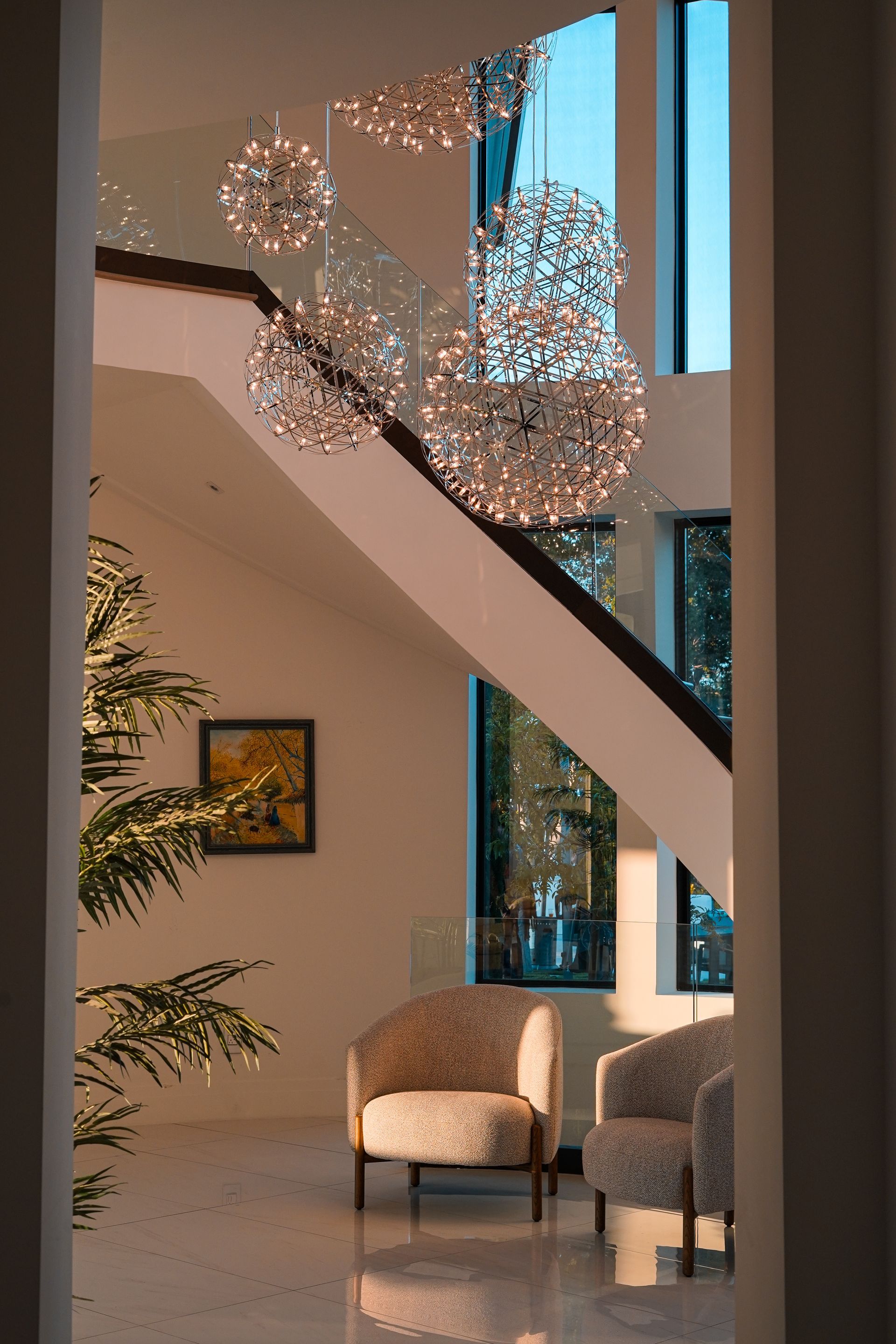 A living room with a staircase , chairs , and a chandelier hanging from the ceiling.