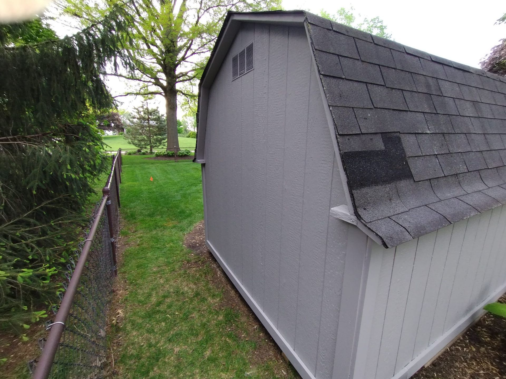 Gray shed with black shingle roof, next to a chain-link fence and green grass.