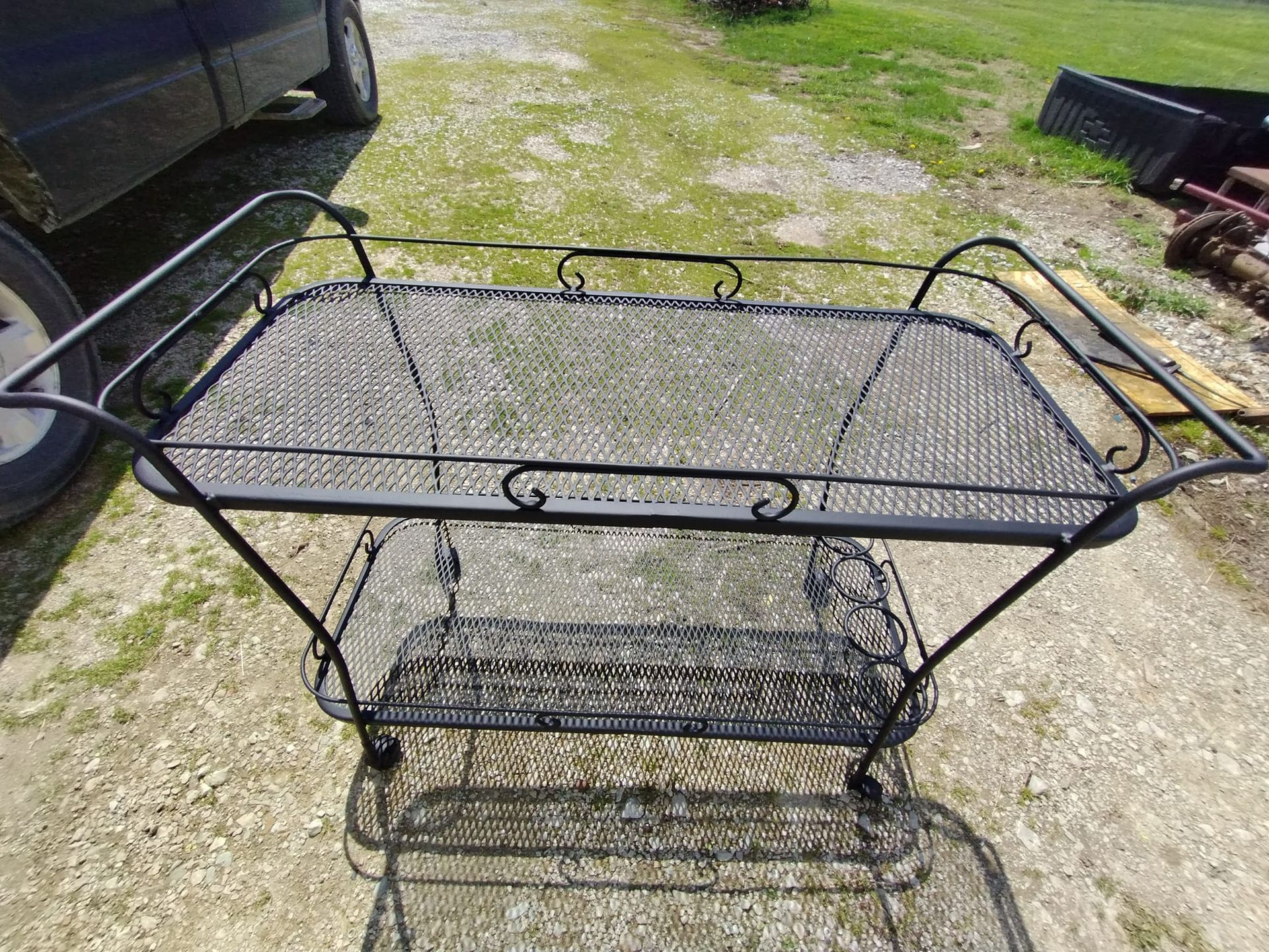 Black metal, two-tiered serving cart with wire mesh shelves and handles outdoors.