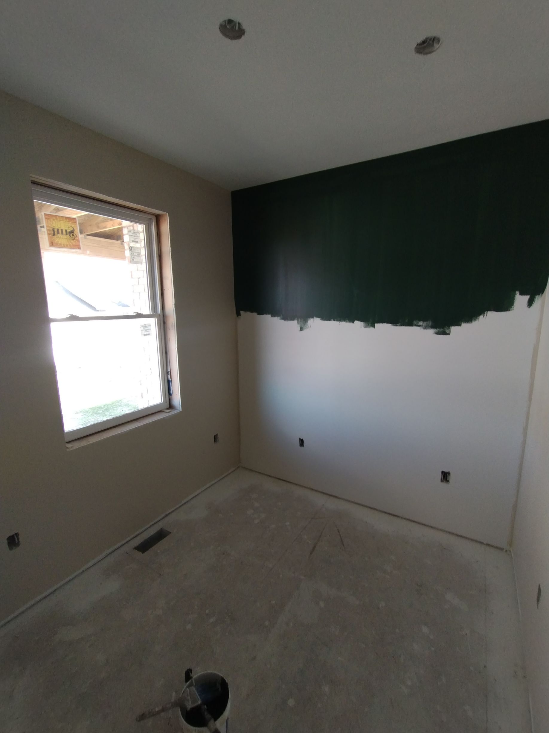 Room with unfinished walls, one painted dark green at top, a window and bare concrete floor.