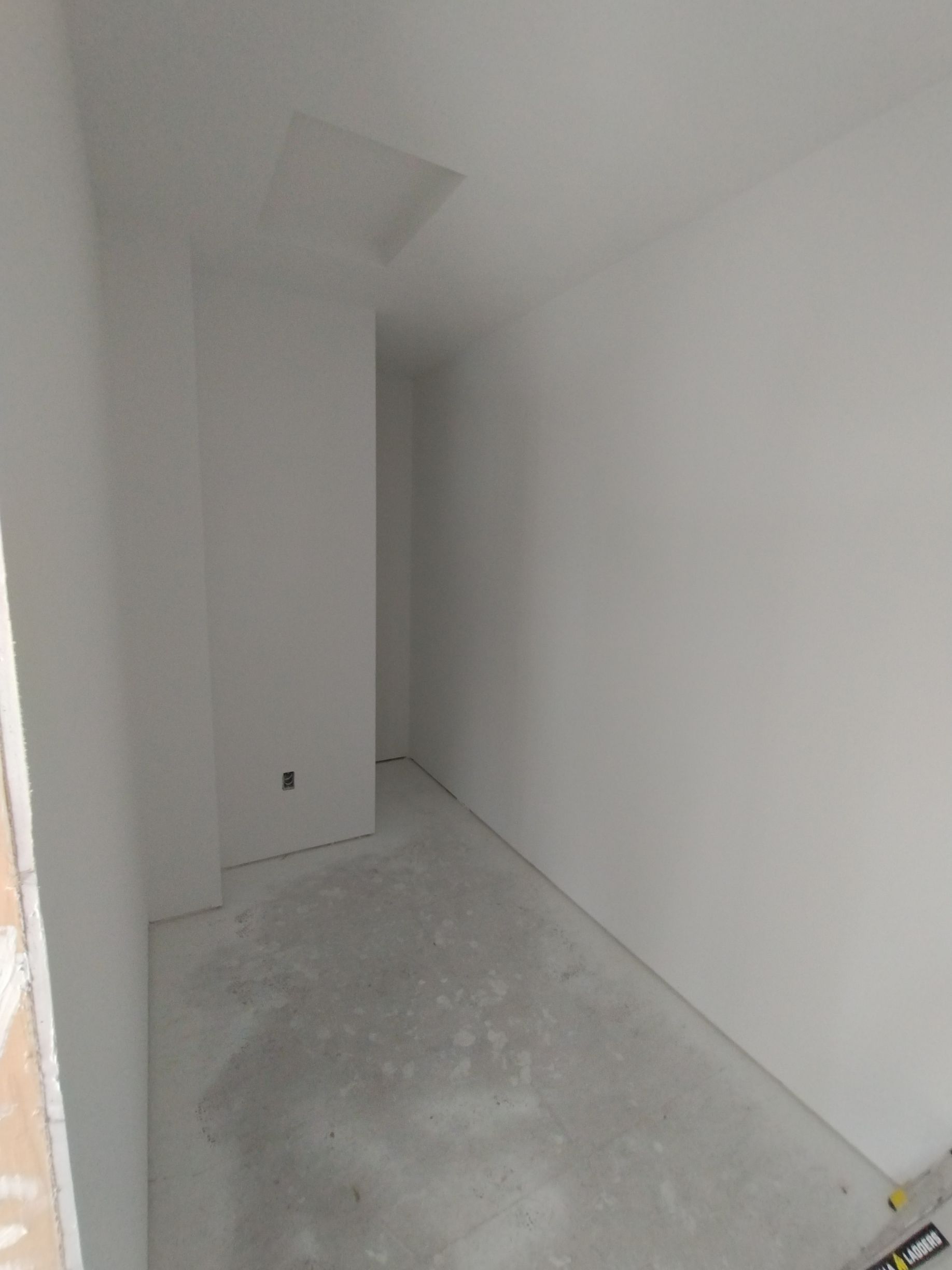 A small, empty room with white walls, concrete floor, and a rectangular opening in the ceiling.