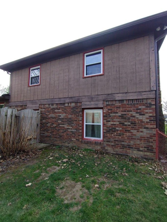 Two-story house with brown siding and brick base, three windows, set against a green lawn and a wooden fence.