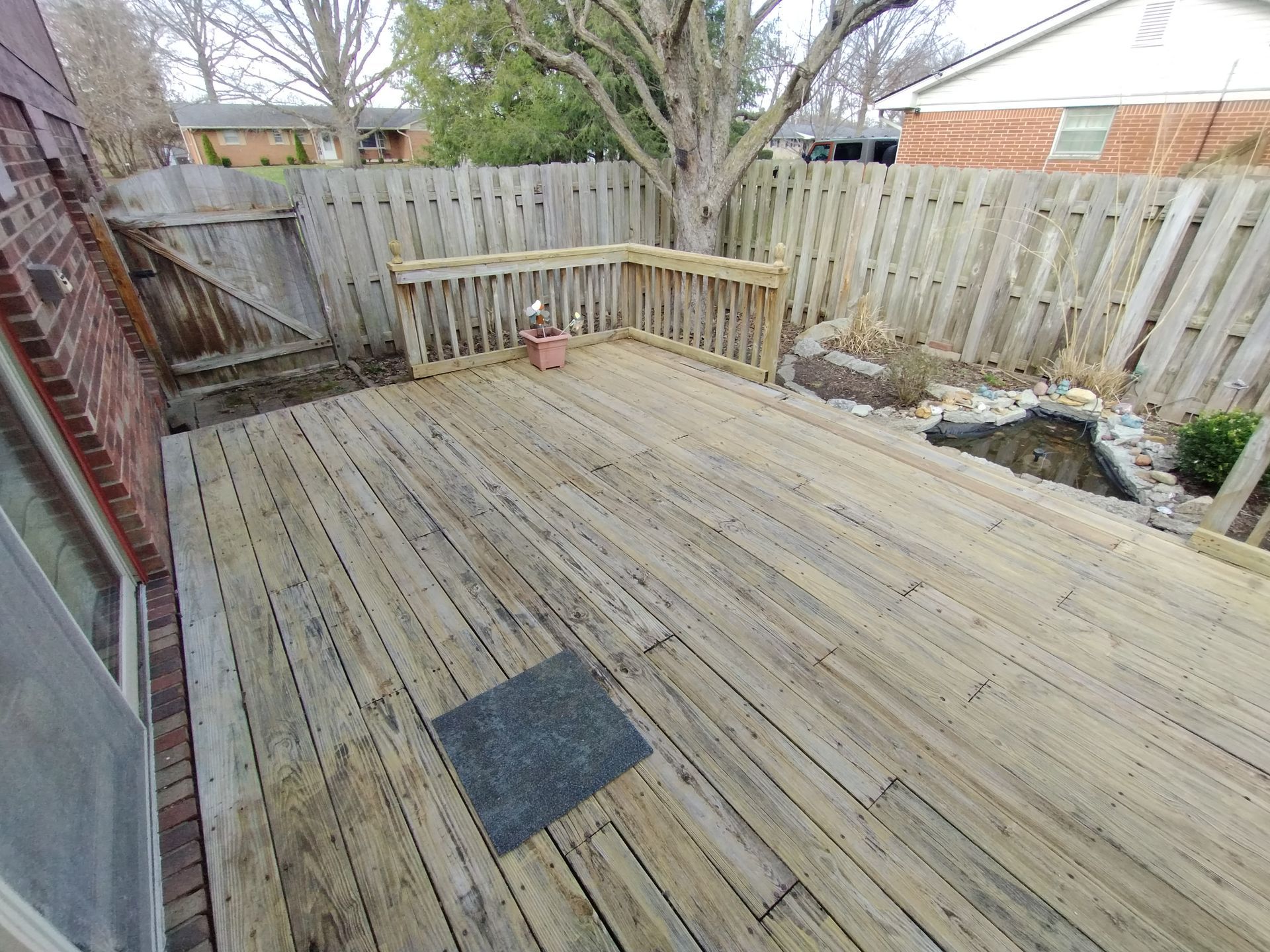 Wooden deck in a backyard, with a fence and pond, and a brick building on the left.