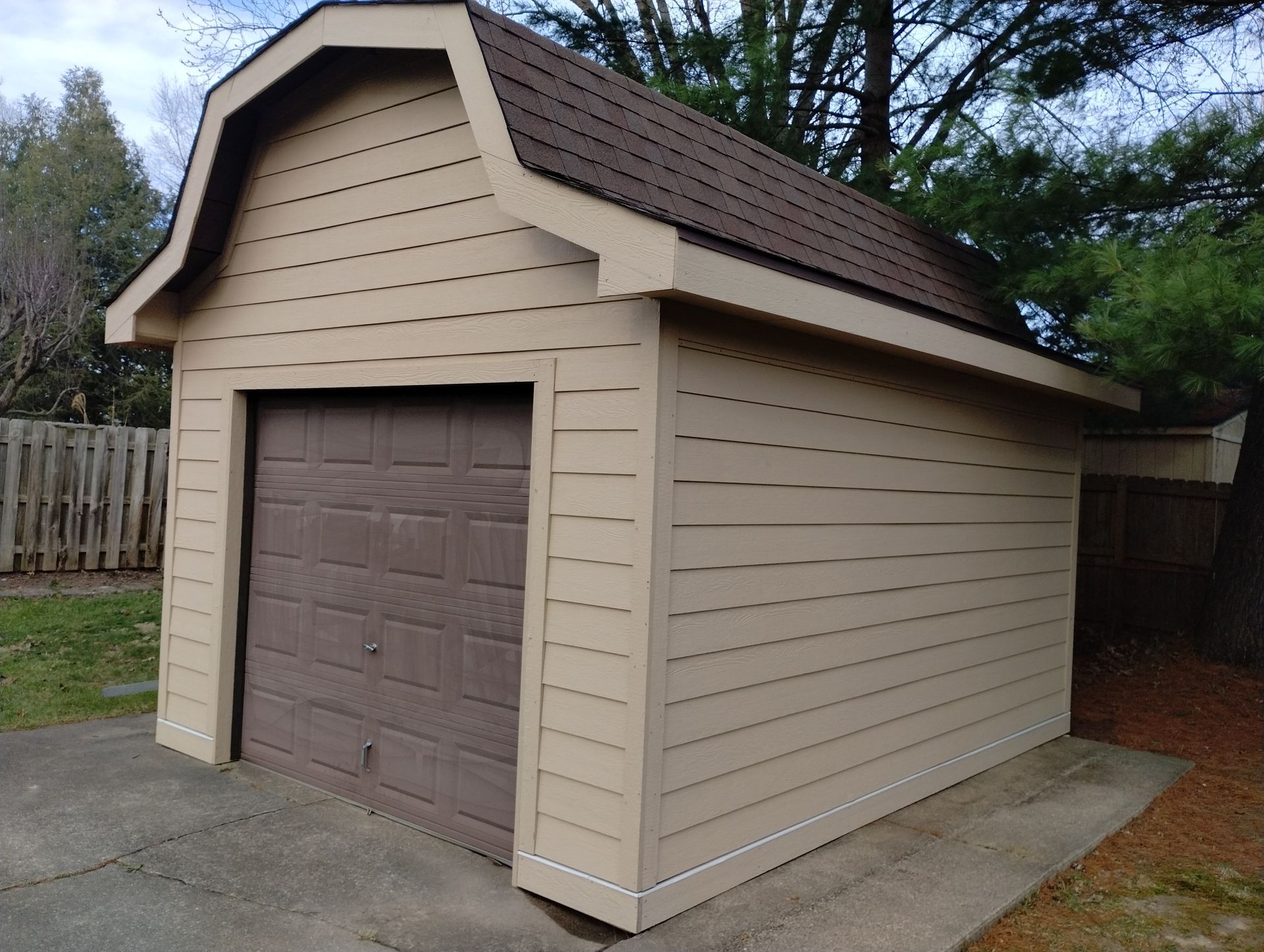 Tan garage with brown roof and door, on a concrete slab next to a wooden fence.