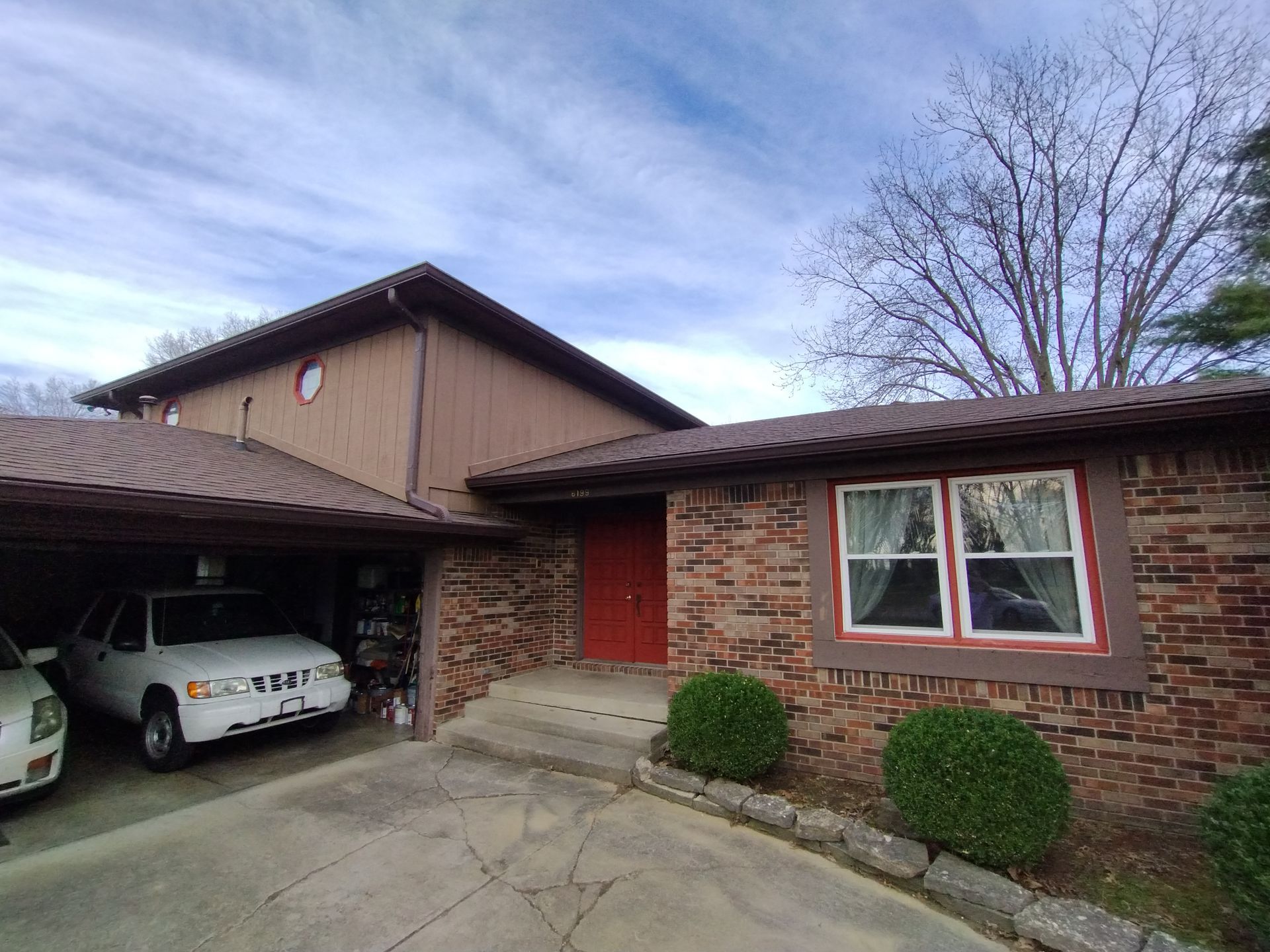 A brown brick house with a car in the carport and red door. Cloudy sky above.