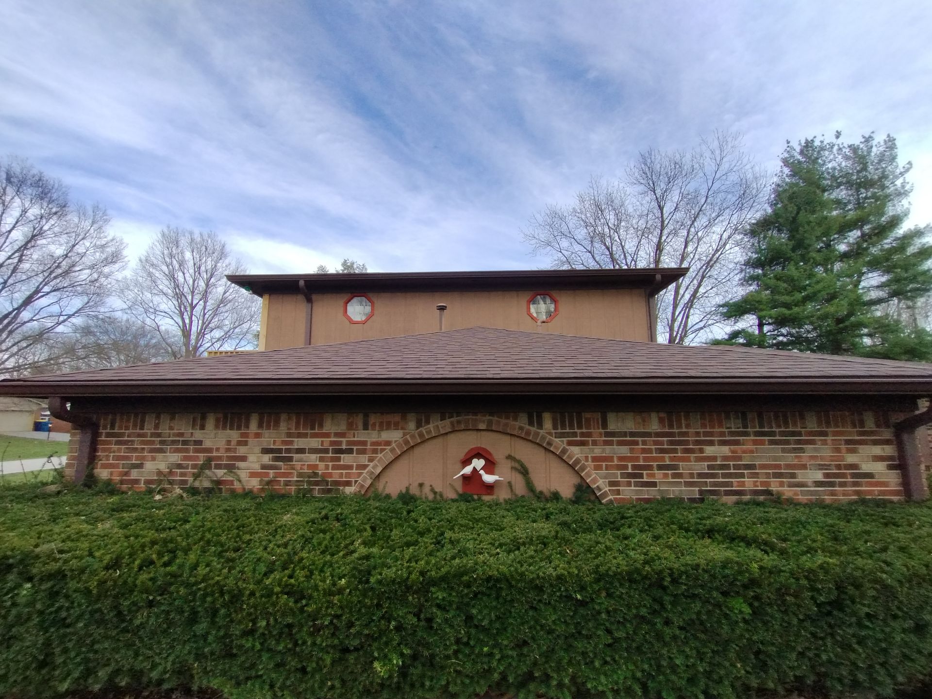 Brick building with brown roof and trim, green hedge in front, wreath above, trees in background, blue sky.