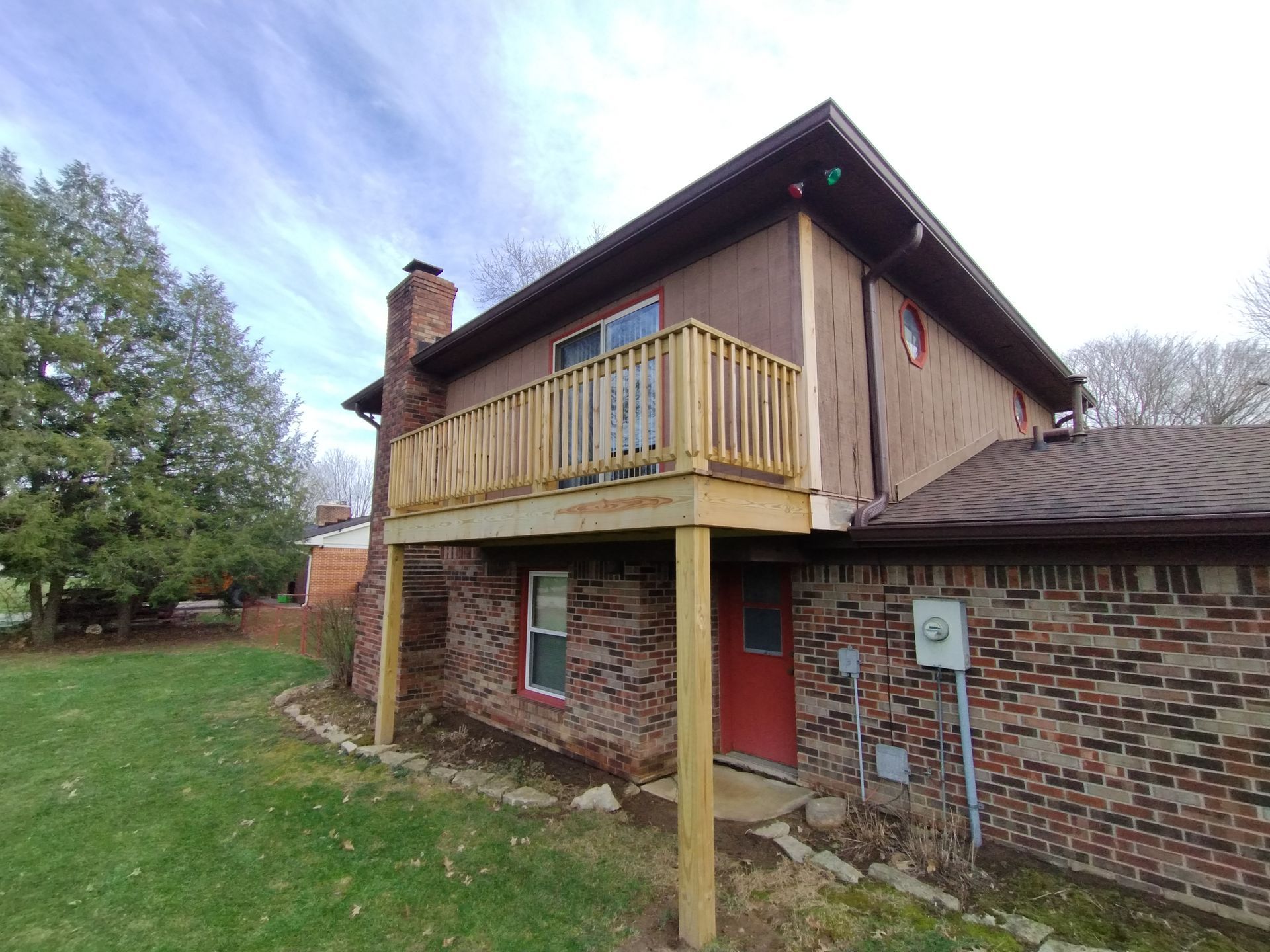 Two-story house with a wooden deck, brown siding, brick, chimney, and red door. Green grass and cloudy sky.