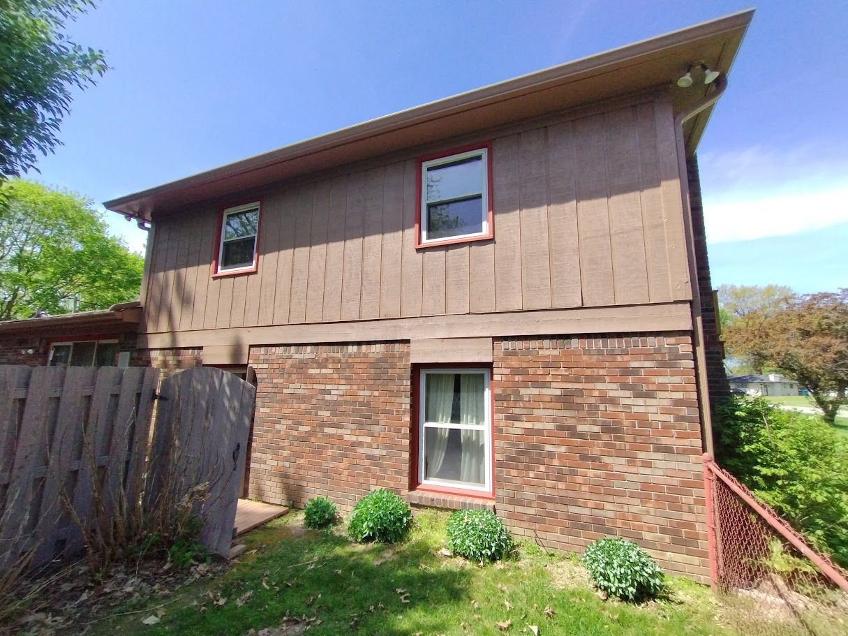 Two-story home with brown siding above brick. Windows have red trim. Wooden fence and green grass in front.