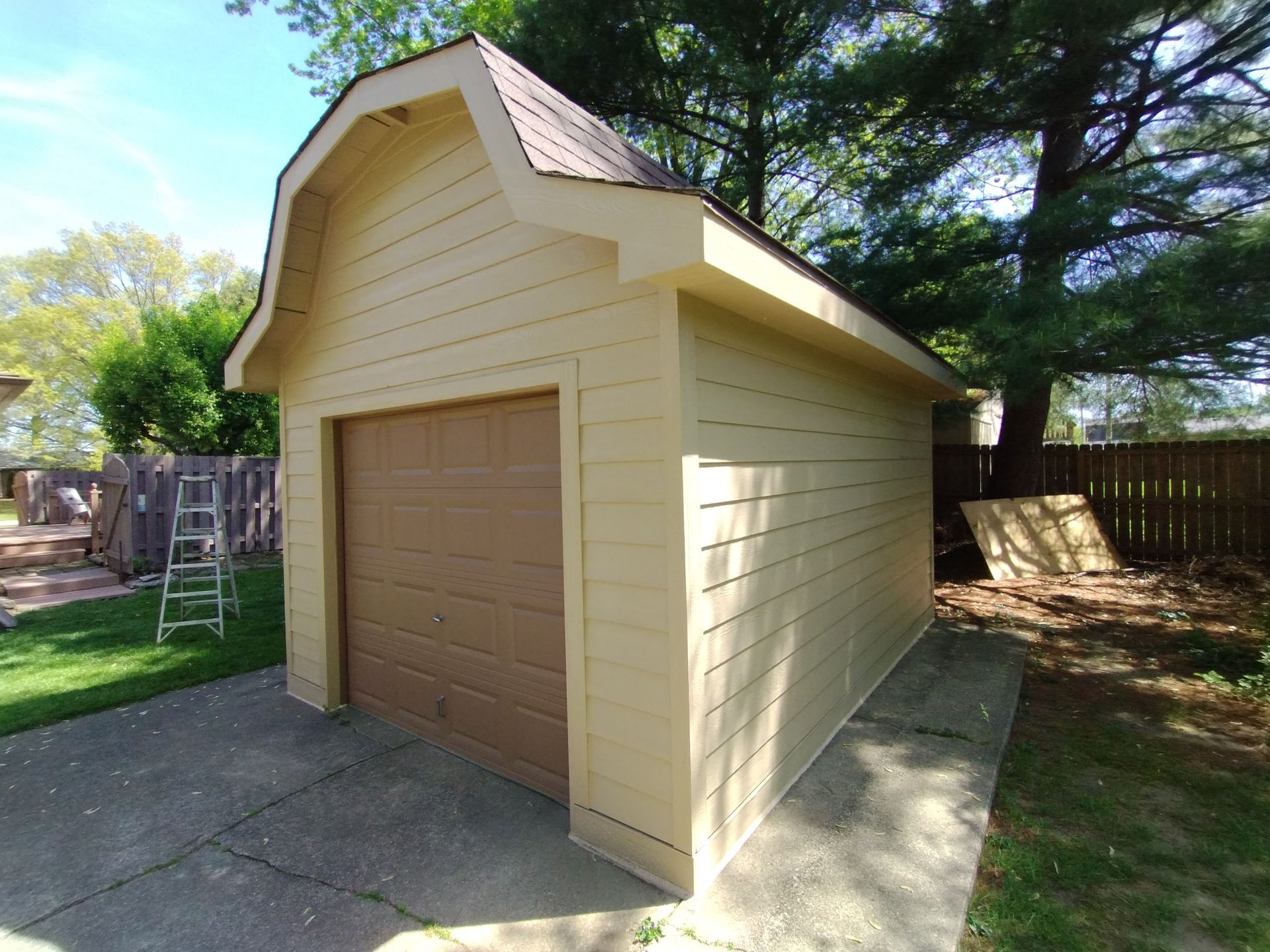 Tan garage with brown door and roof in a backyard setting with green grass and trees.