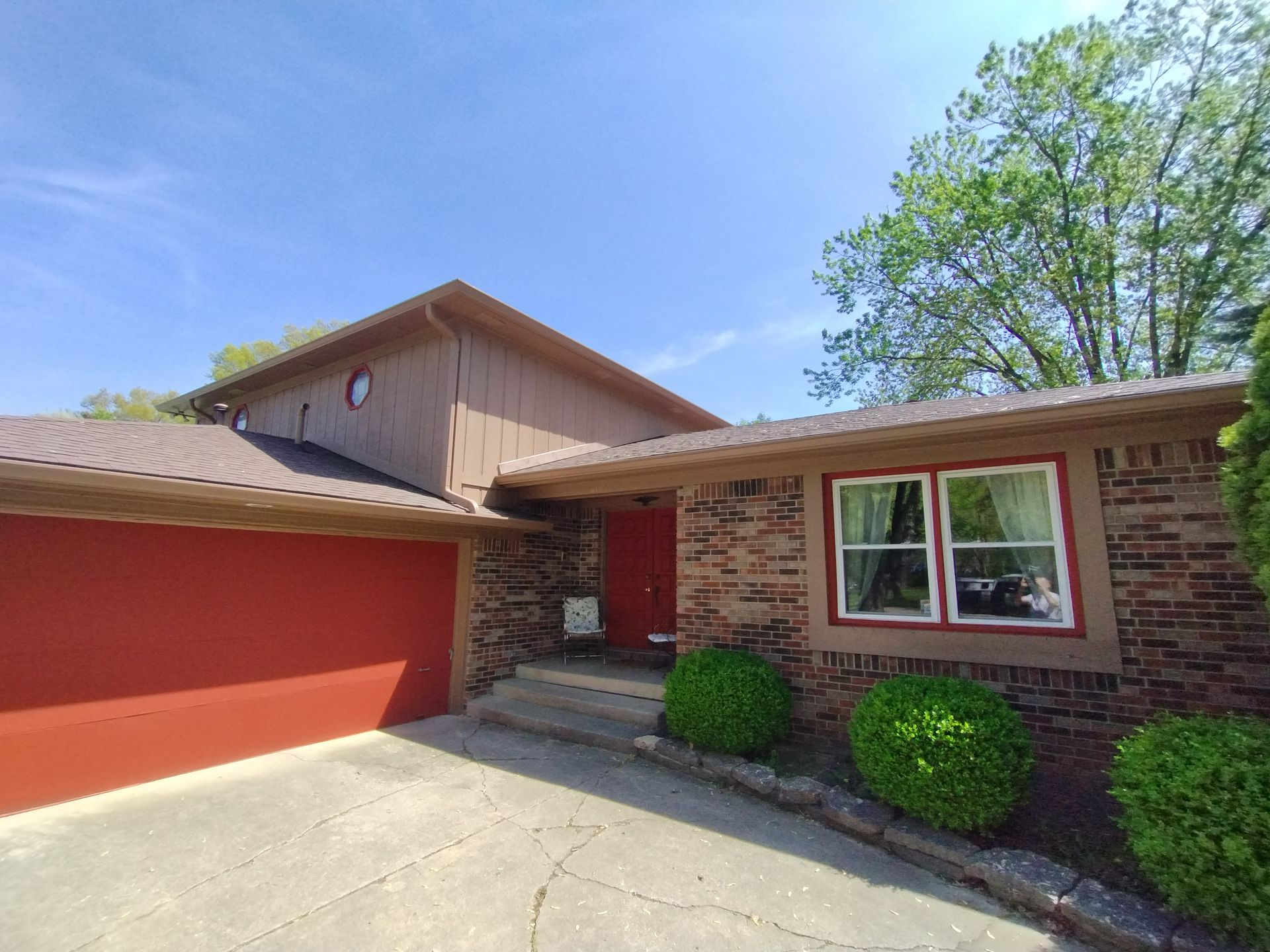 Brown brick house with red accents, garage door, and window frames under a blue sky.