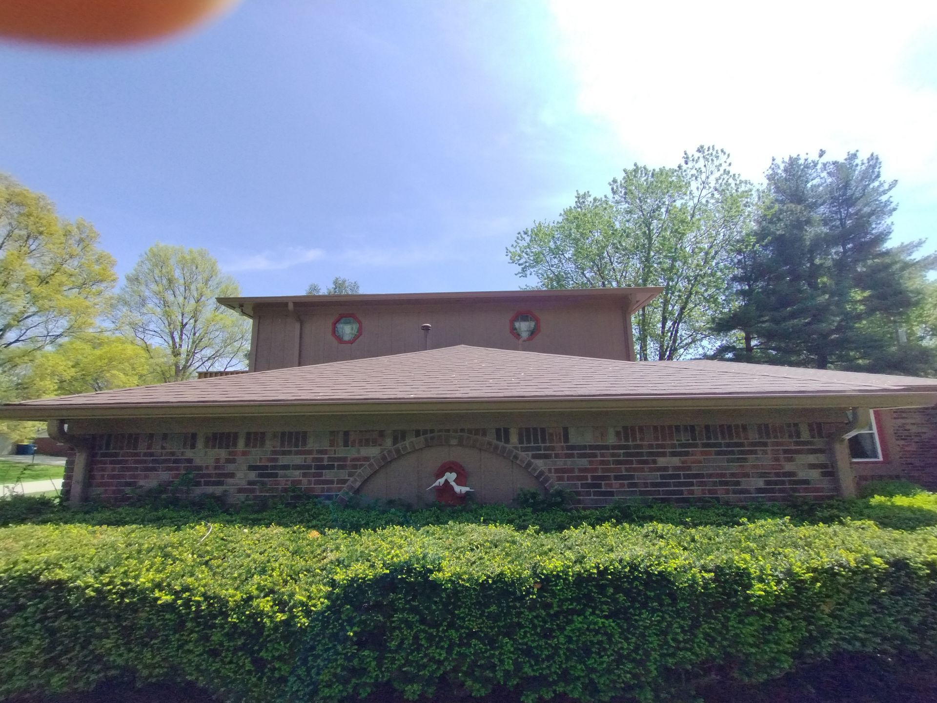 Low-angle view of a brick and brown roof house with circular windows, trees, and a blue sky background.