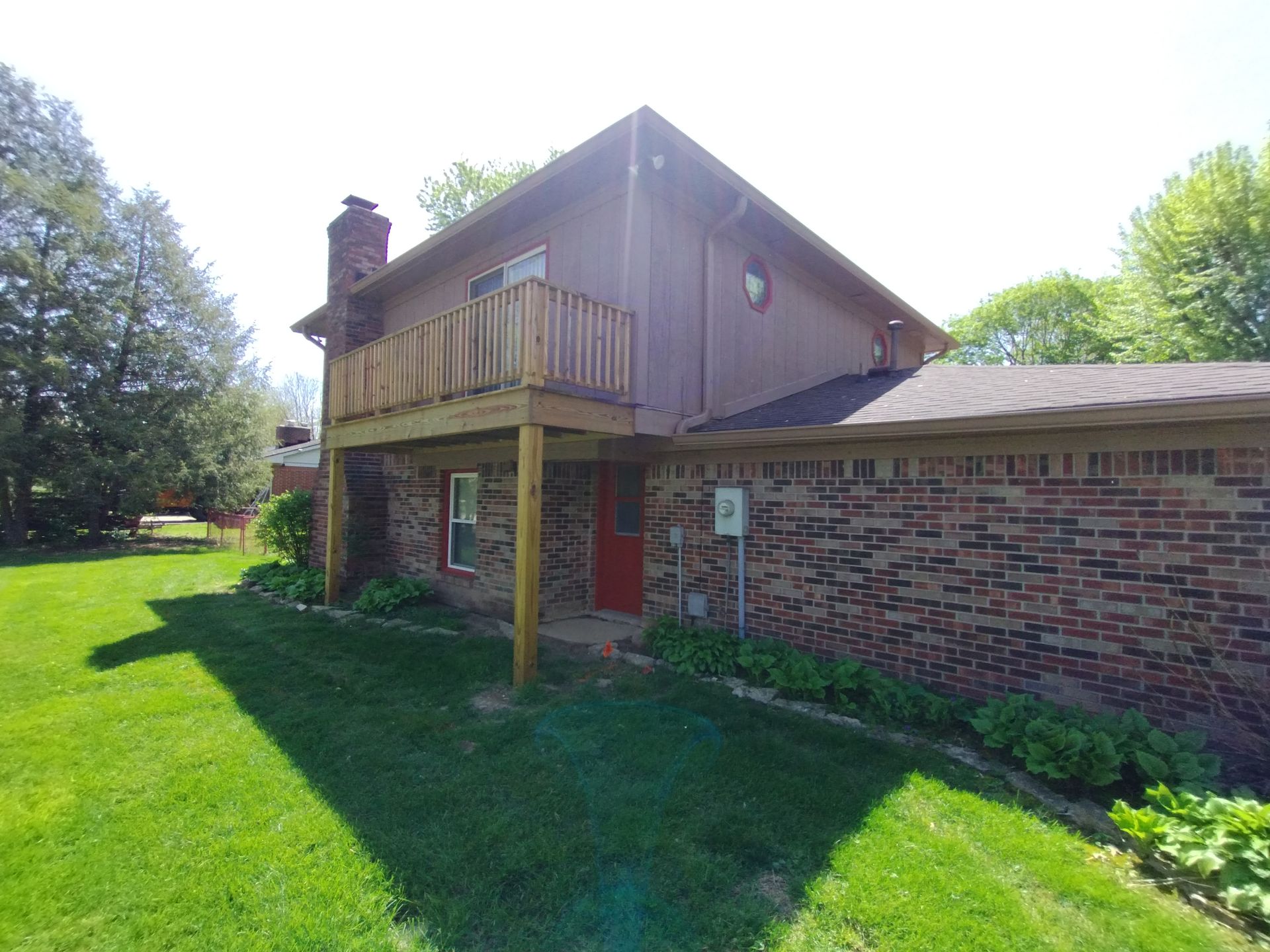 Brown two-story house with a wooden deck. Brick wall and green lawn.