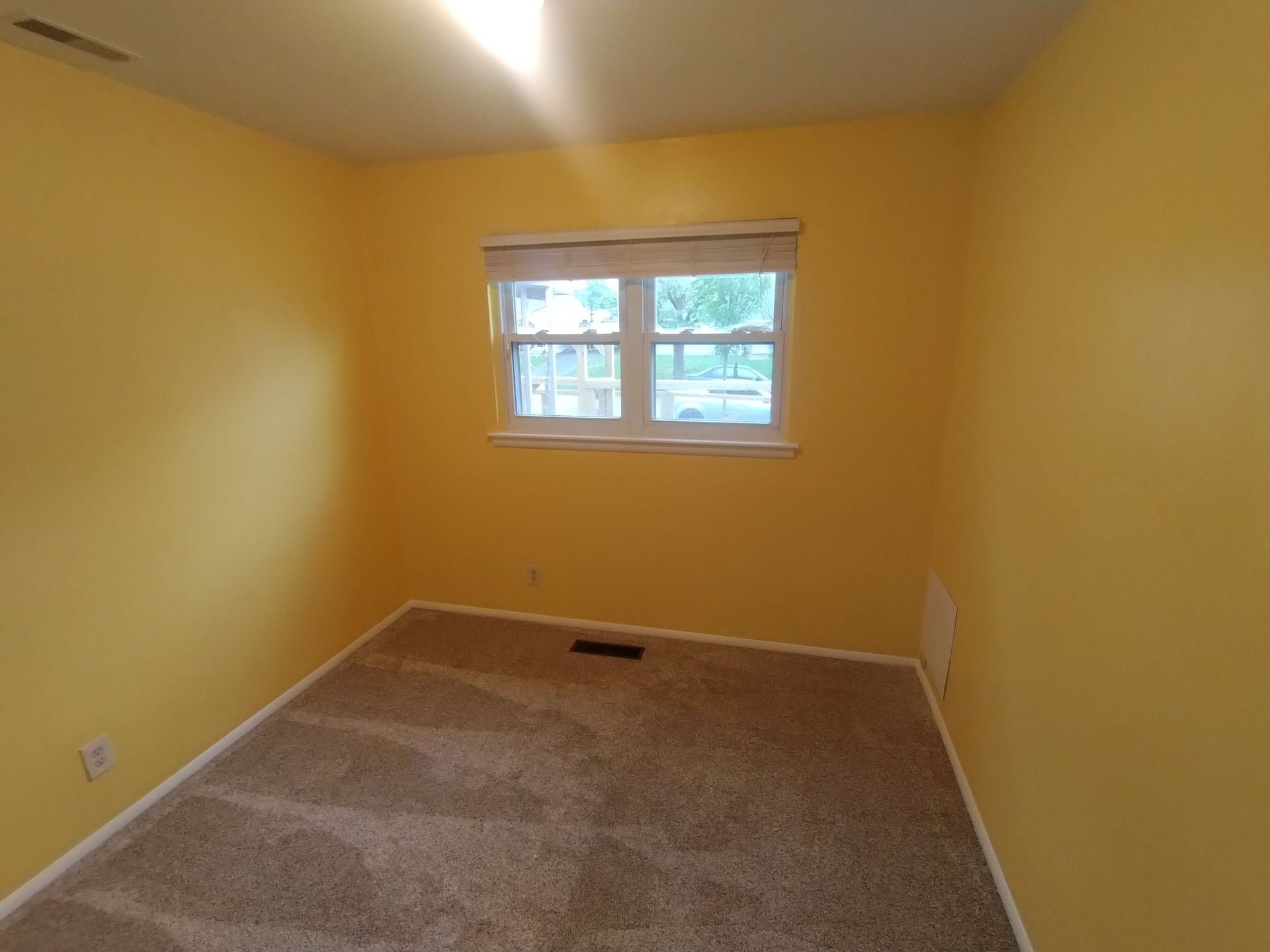 Yellow-walled room with beige carpet, small window with blinds, and electrical outlets.