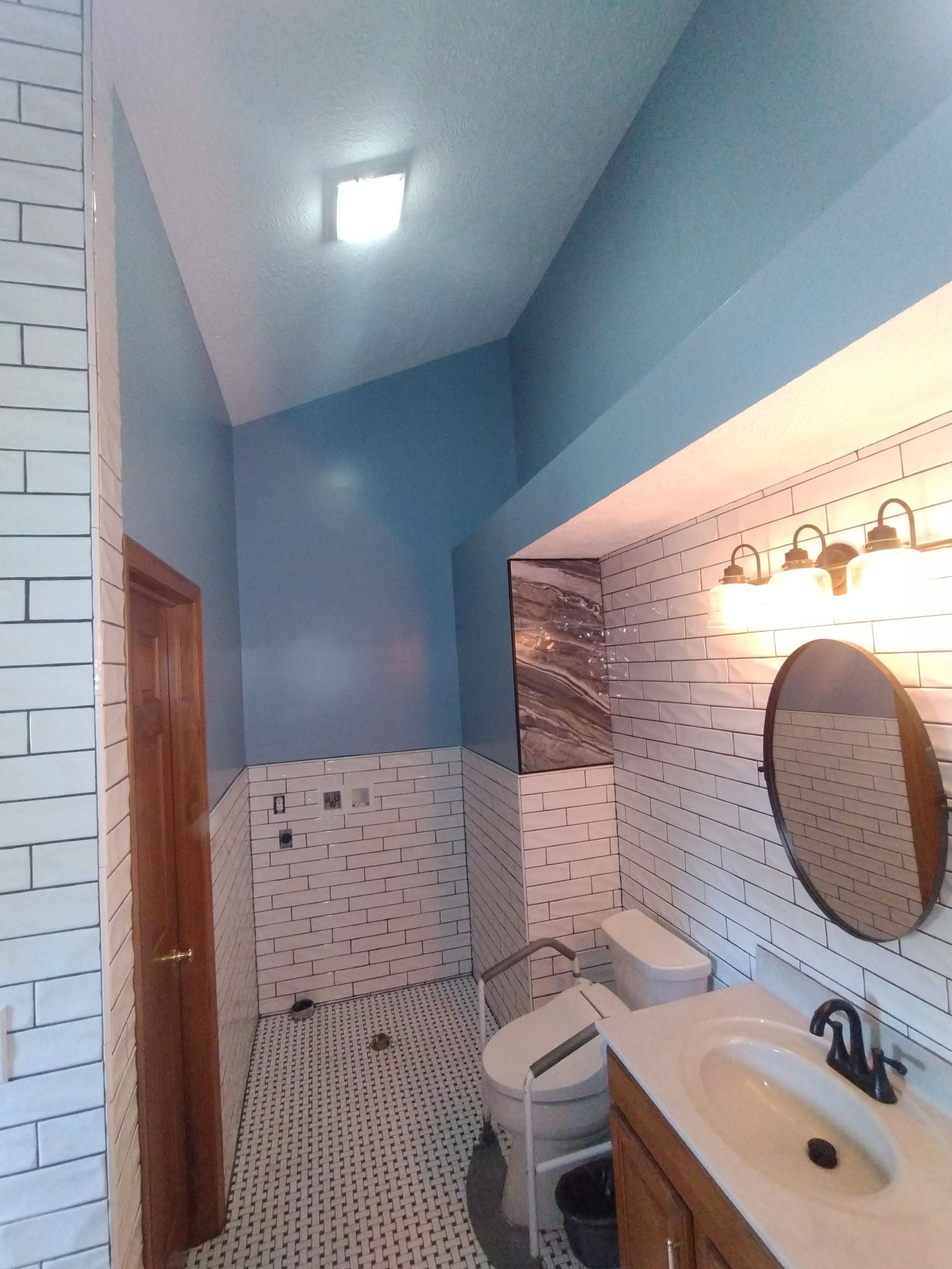 Bathroom with white tile, blue walls, wooden door, vanity, toilet, and round mirror.