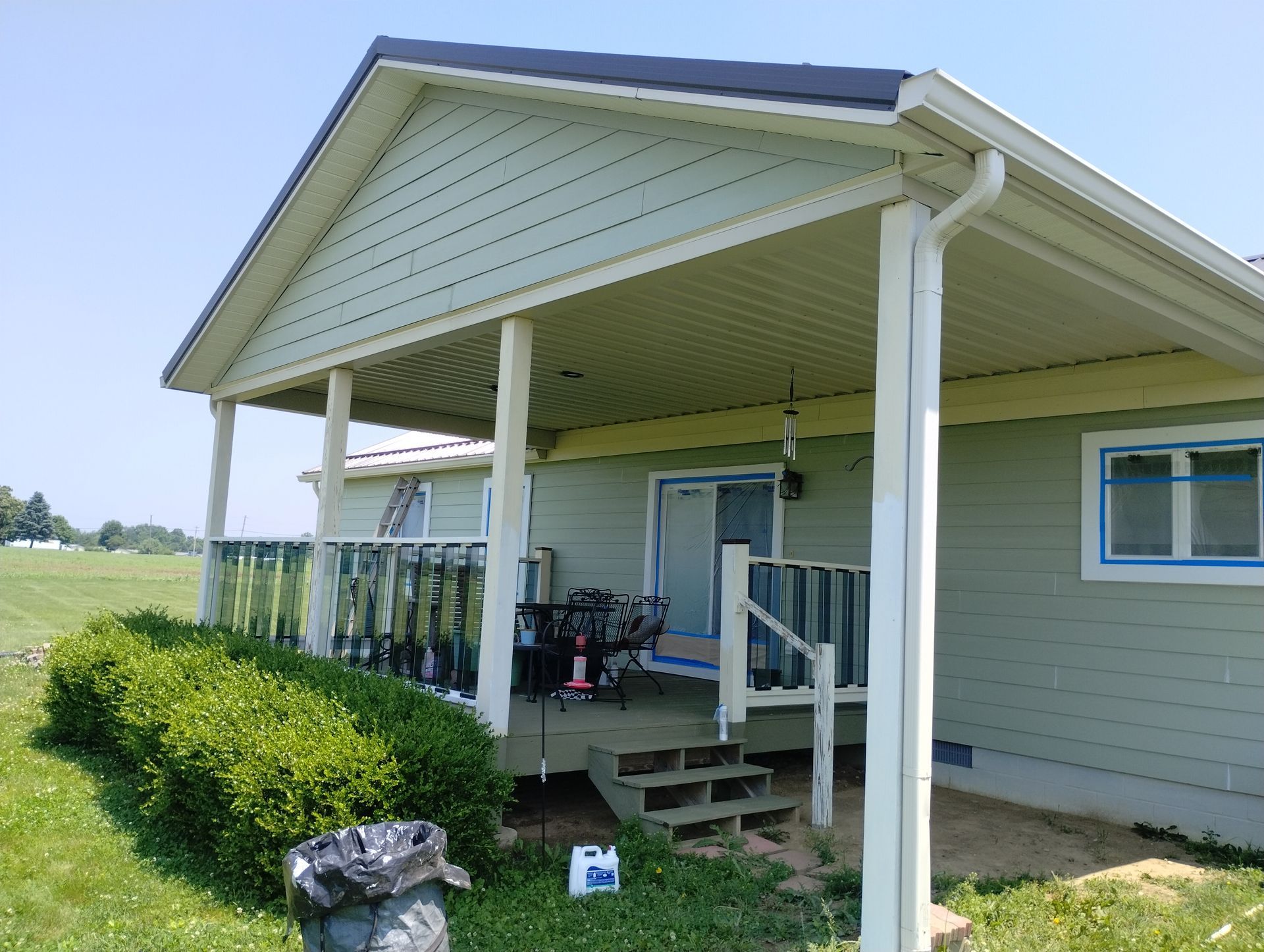 A green house with a covered porch, railing, and small flight of stairs. Includes a trash can and a field.