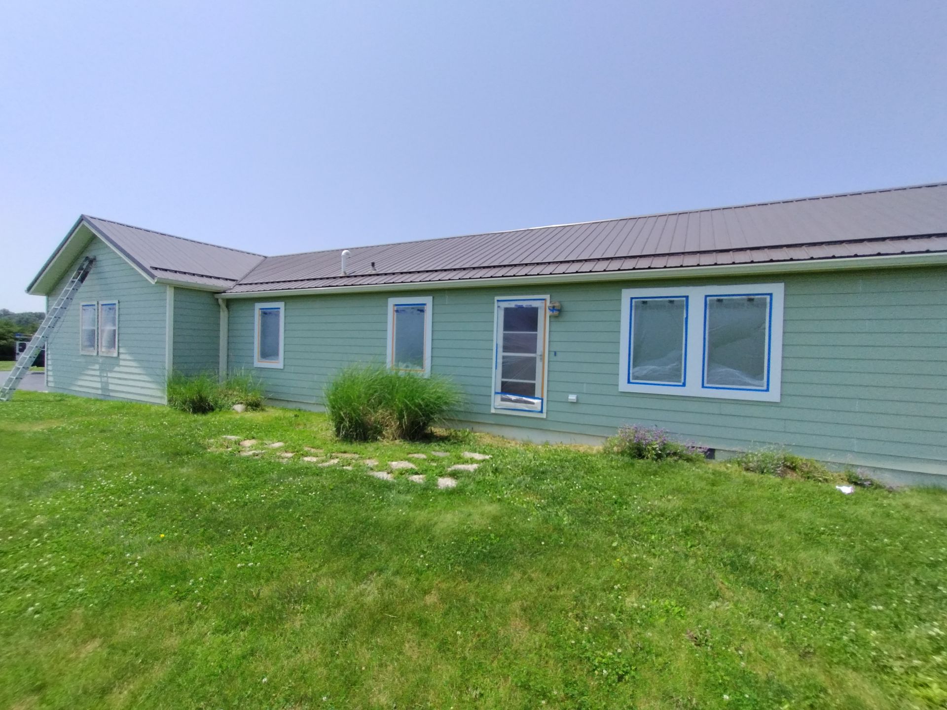 Green house with dark roof, windows taped for painting, ladder, and yard under a blue sky.