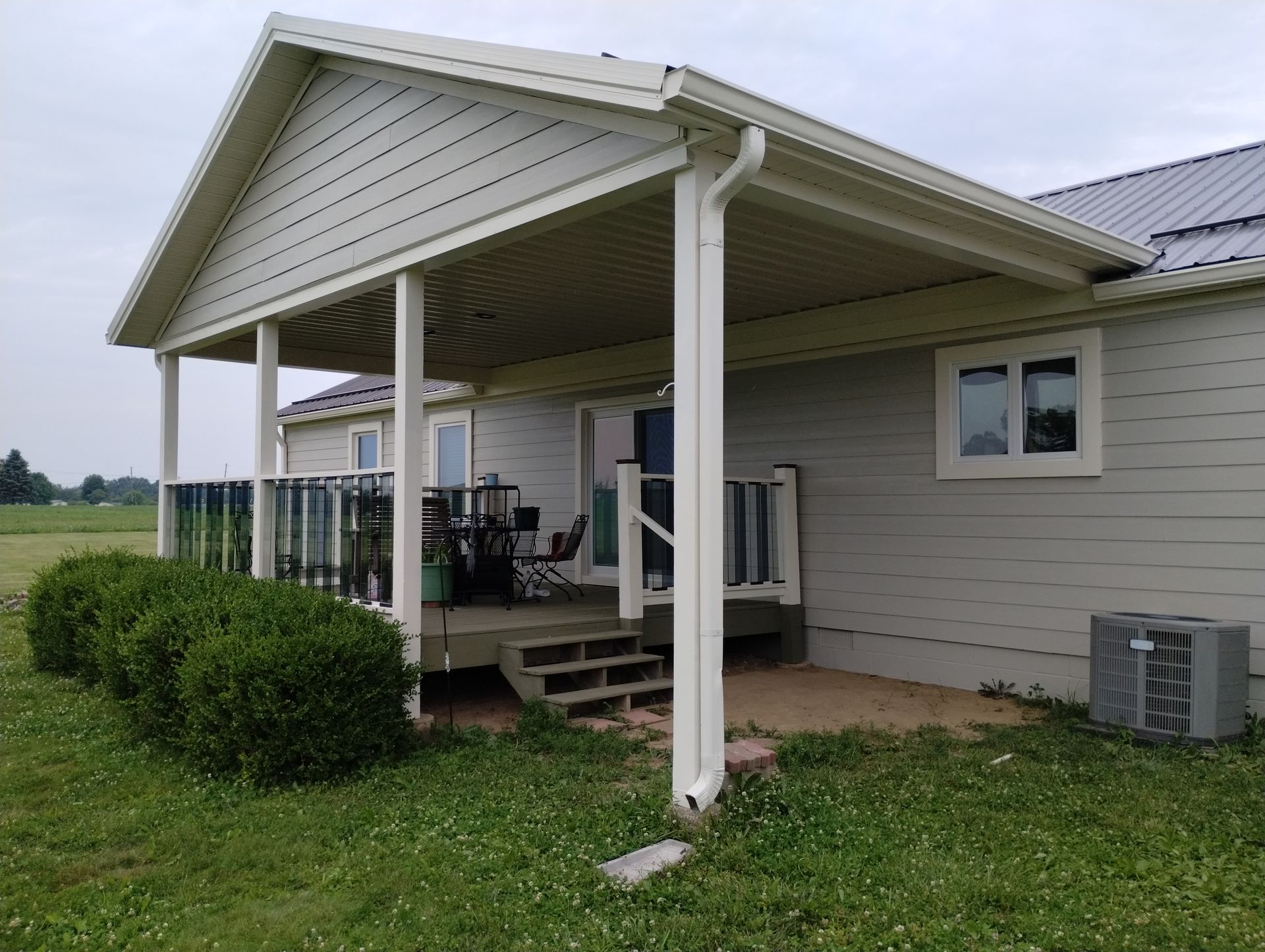 Beige-sided house with a covered porch, wooden deck, and bushes, set in a grassy yard.