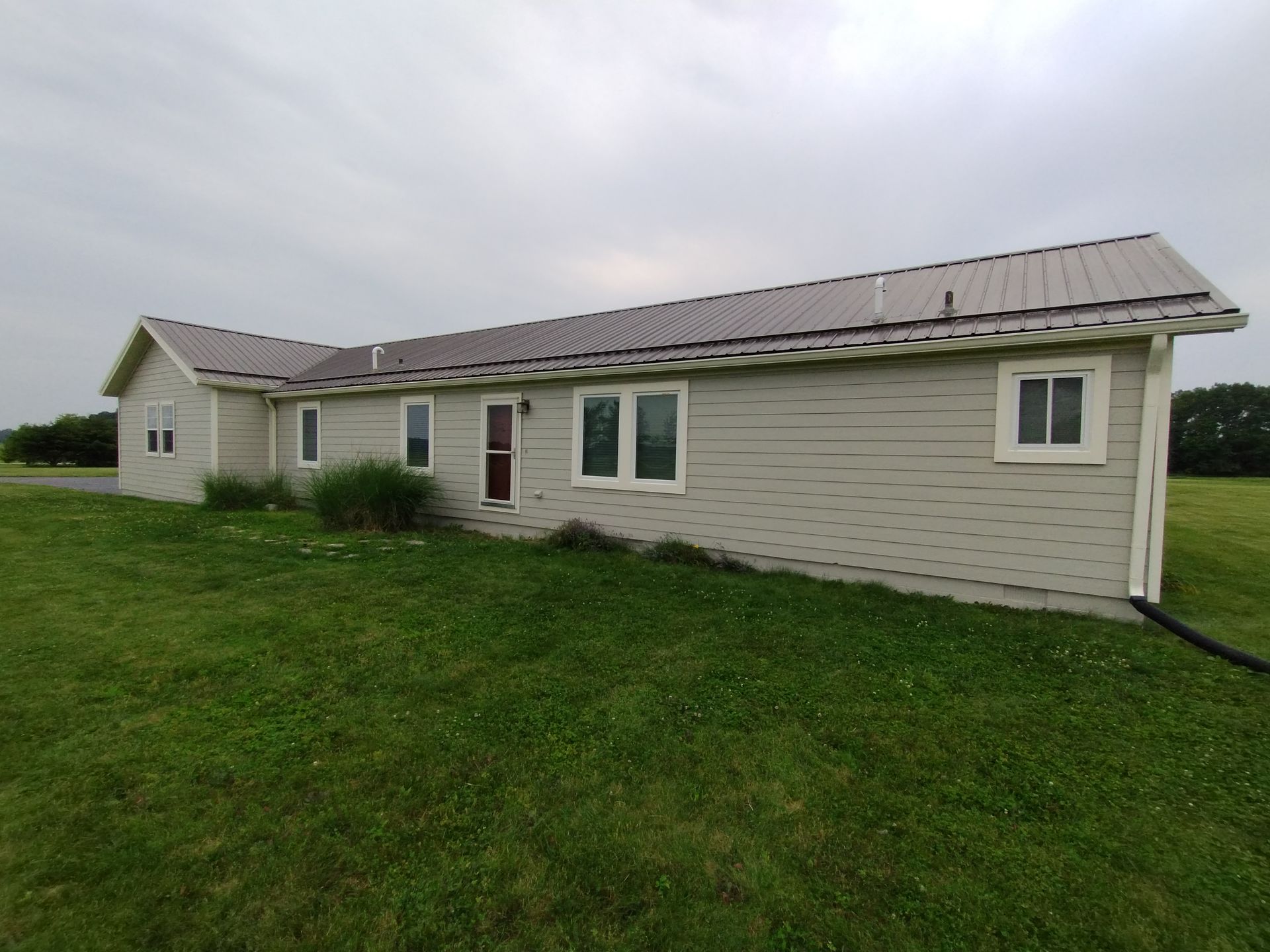 Beige ranch house with metal roof, windows, and green lawn under a cloudy sky.