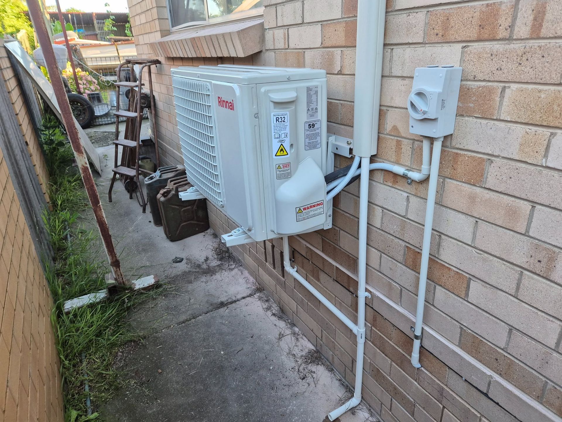 Outdoor air conditioner unit and pipes mounted beside a brick house wall in a narrow side yard