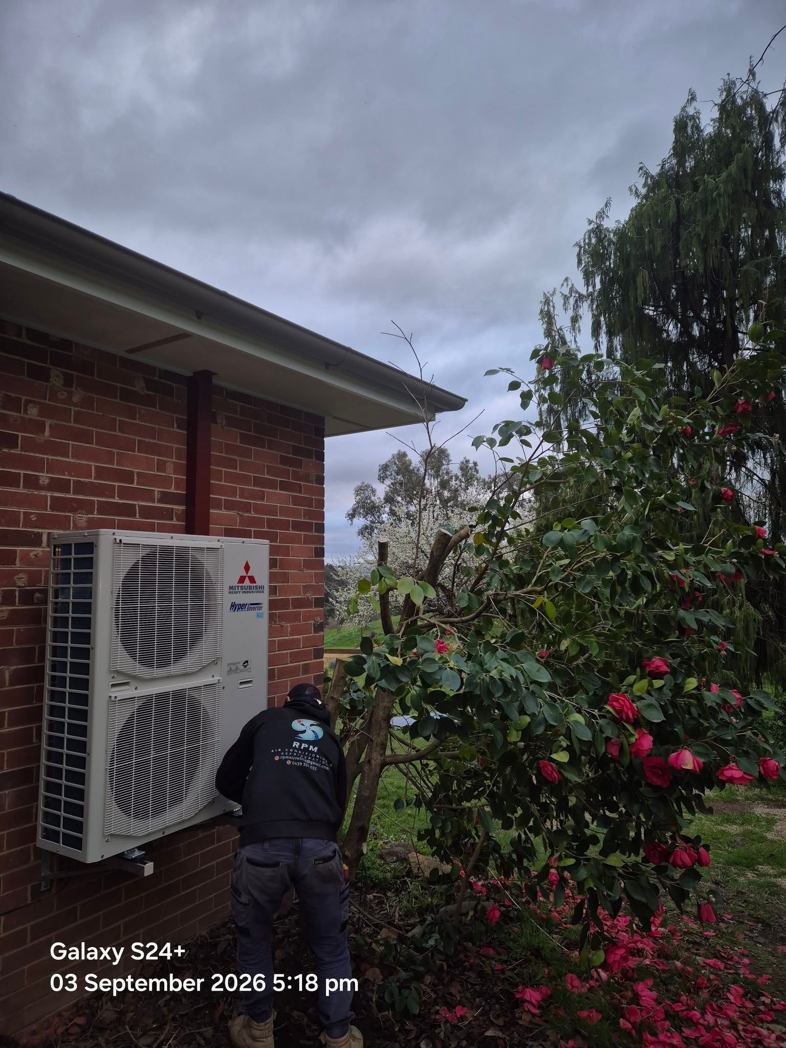 Outdoor air conditioner unit beside a brick house and lush flowering garden under a cloudy sky