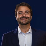 Homem barbudo sorrindo, vestindo uma camisa polo azul-clara. Contra uma parede cinza texturizada.