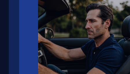 Homem dirigindo um carro conversível, perfil lateral. Camisa azul, concentrado na estrada à frente, ao ar livre.