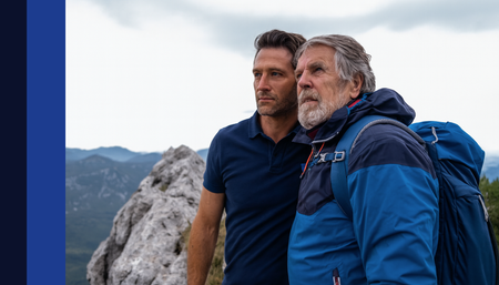 Dois homens no topo de uma montanha, um mais velho com uma mochila, contemplando uma paisagem nublada.