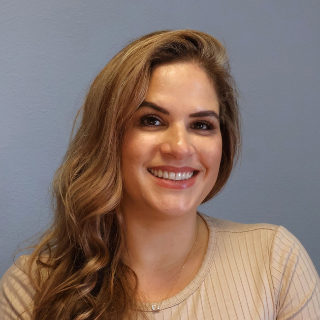 Smiling person with long, wavy brown hair, wearing a ribbed beige top, set against a plain gray background.