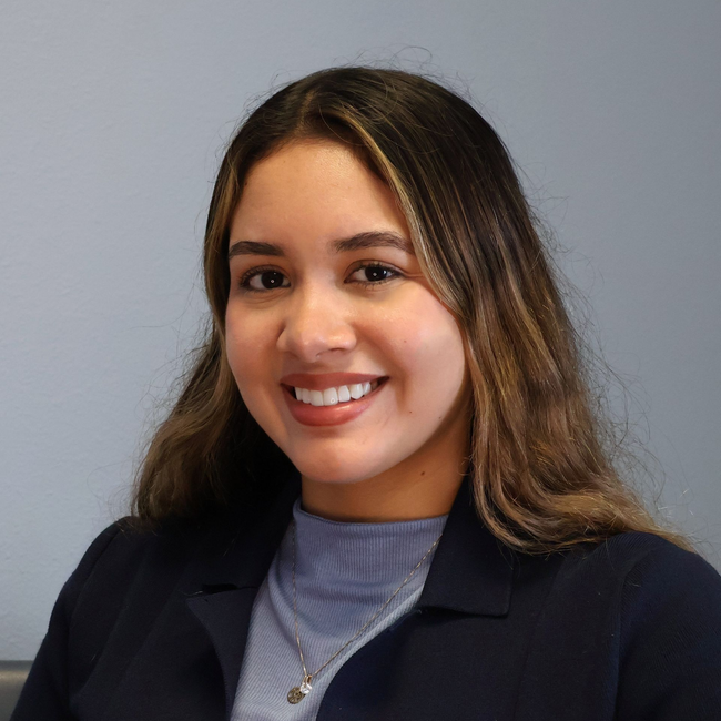 A professional headshot of a person with long, wavy brown hair, smiling, wearing a blue shirt under a dark blazer.