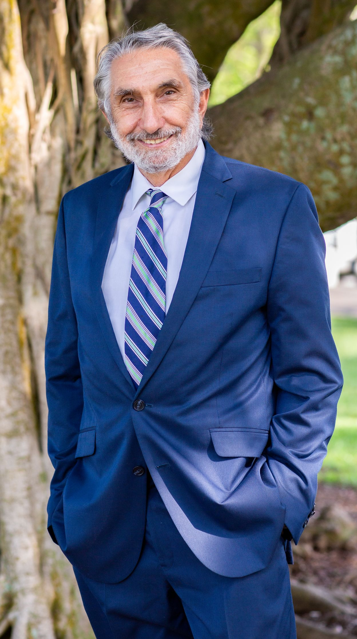 A man in a navy blue suit and a patterned blue tie stands smiling in front of a tree.