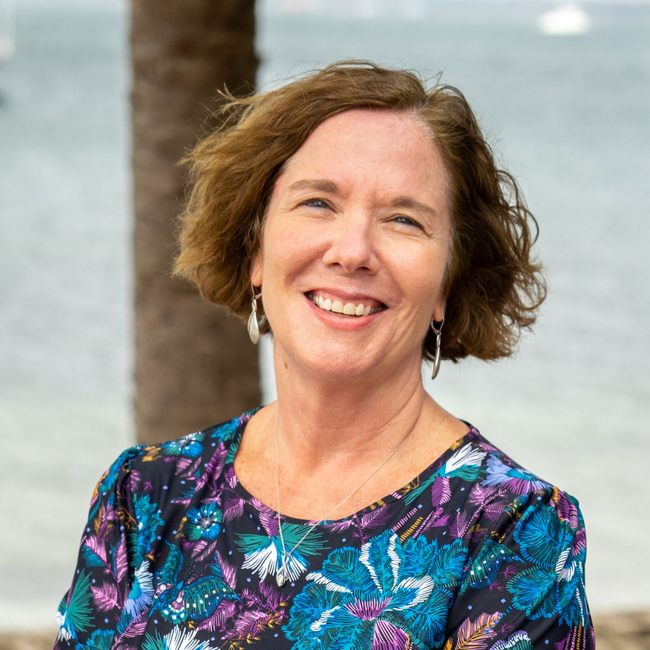 A smiling person with shoulder-length hair wears a floral top and silver earrings, posed before a beach backdrop.