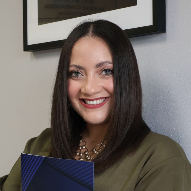 A smiling person with dark hair and a gold beaded necklace holds a blue patterned folder in front of a grey wall.