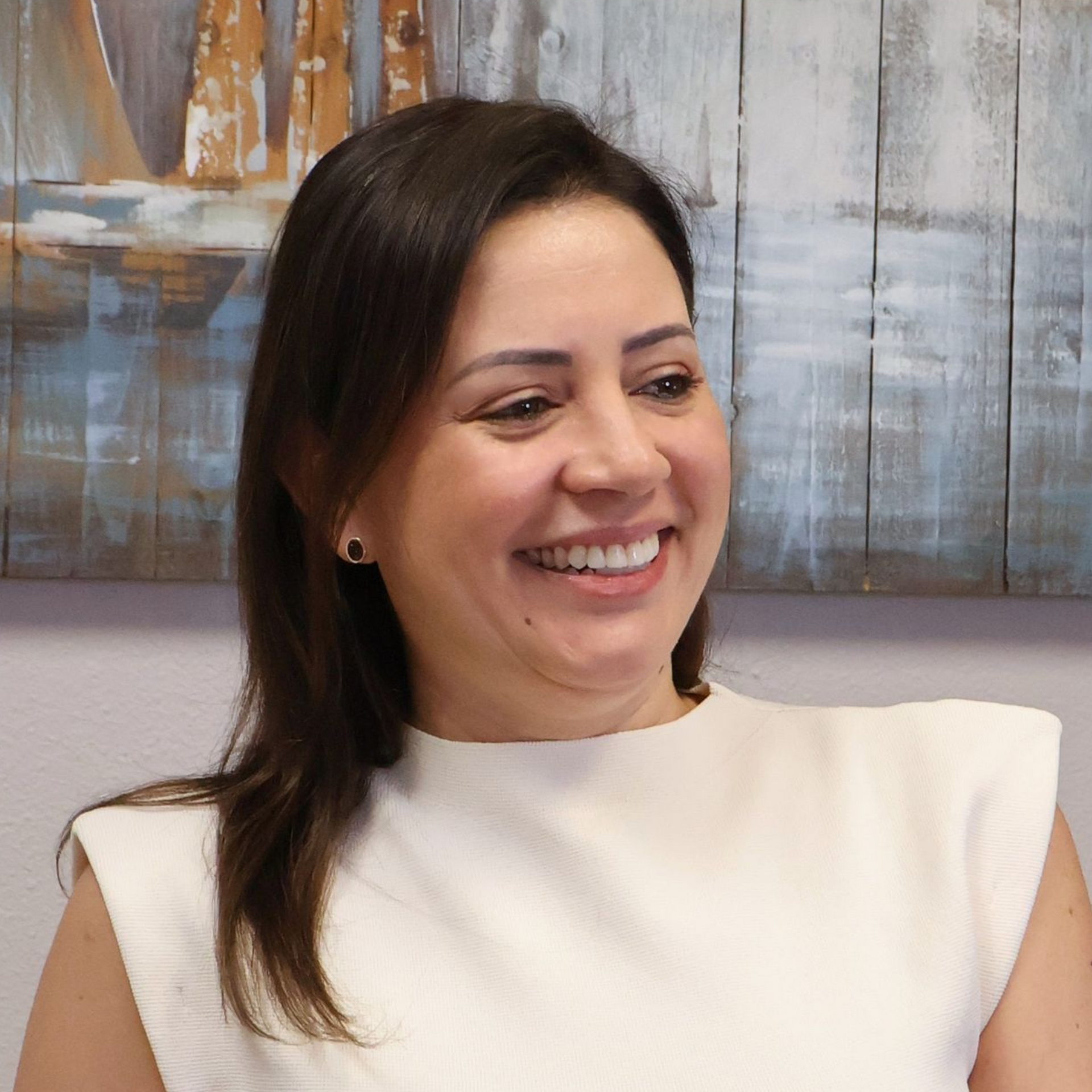 A person with dark hair, wearing a white top, smiles against a background of textured, multi-colored wooden planks.