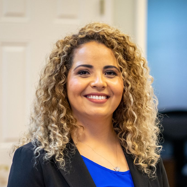 A smiling person with shoulder-length curly blonde hair wearing a blue top under a dark blazer in an office setting.
