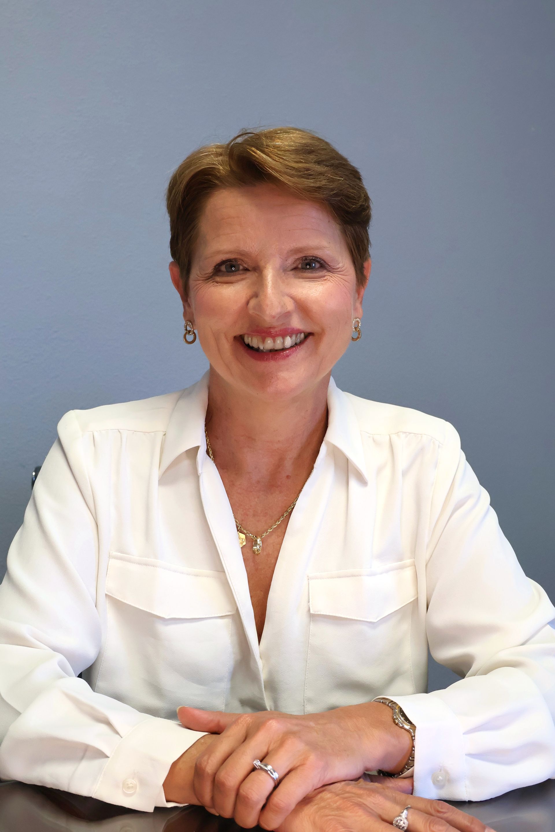 A person with short brown hair wears a white button-down shirt and smiles while resting their hands on a surface.