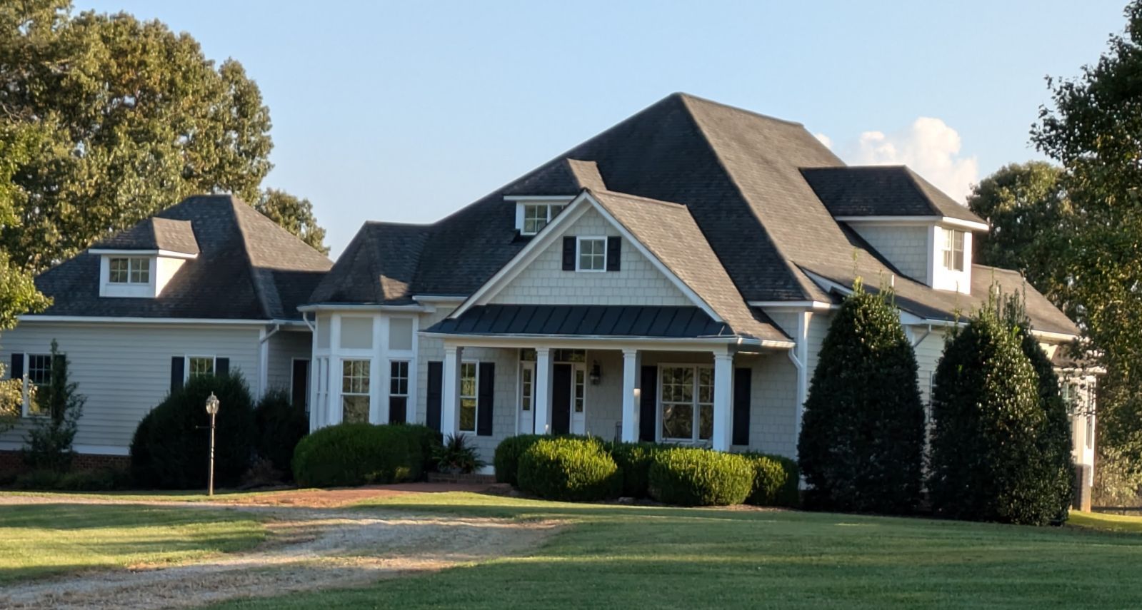 A large, two-story, white house with a dark gray roof, surrounded by green grass and trees under a blue sky.