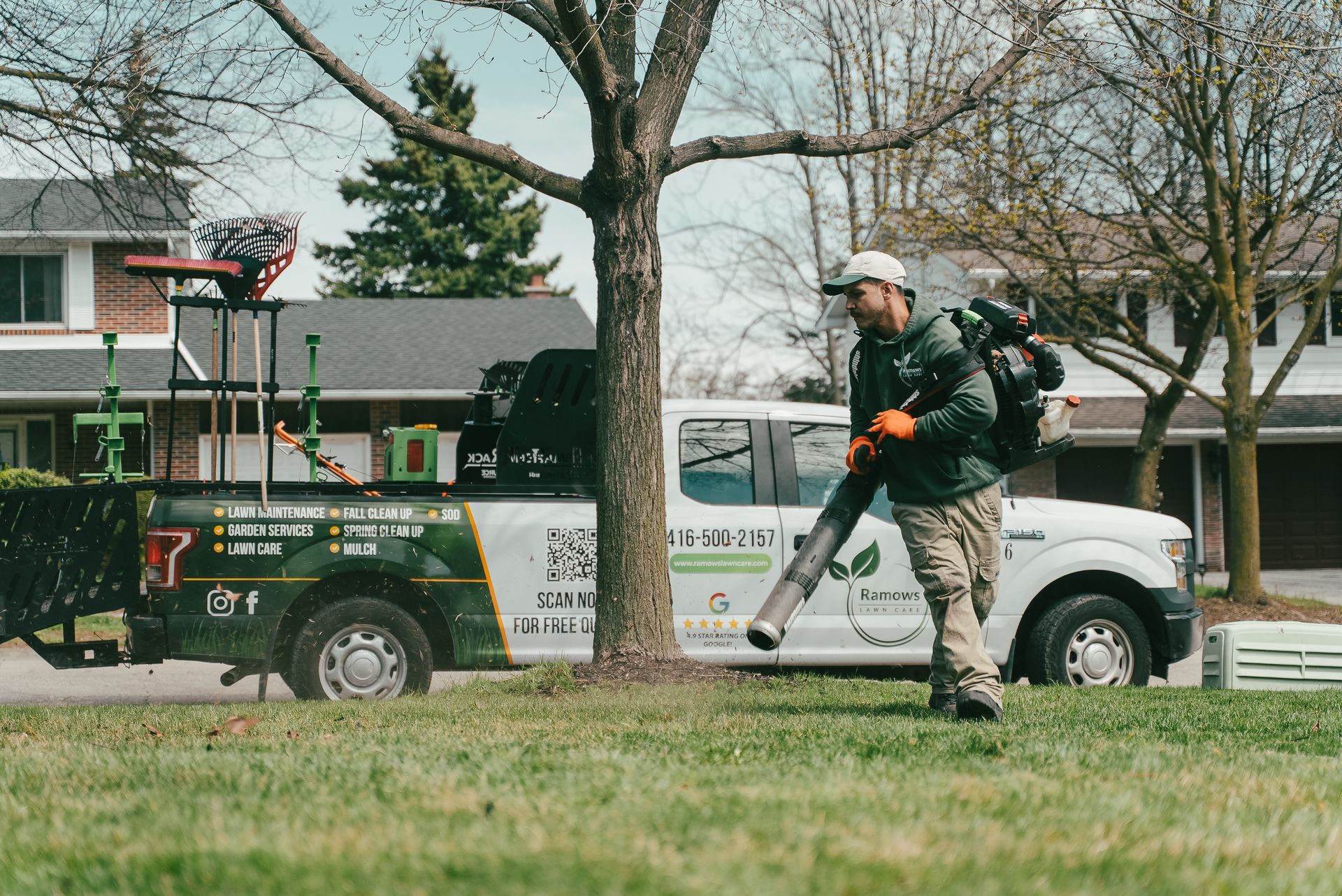 Worker using a leaf blower on a grassy roadside near parked utility trucks and houses