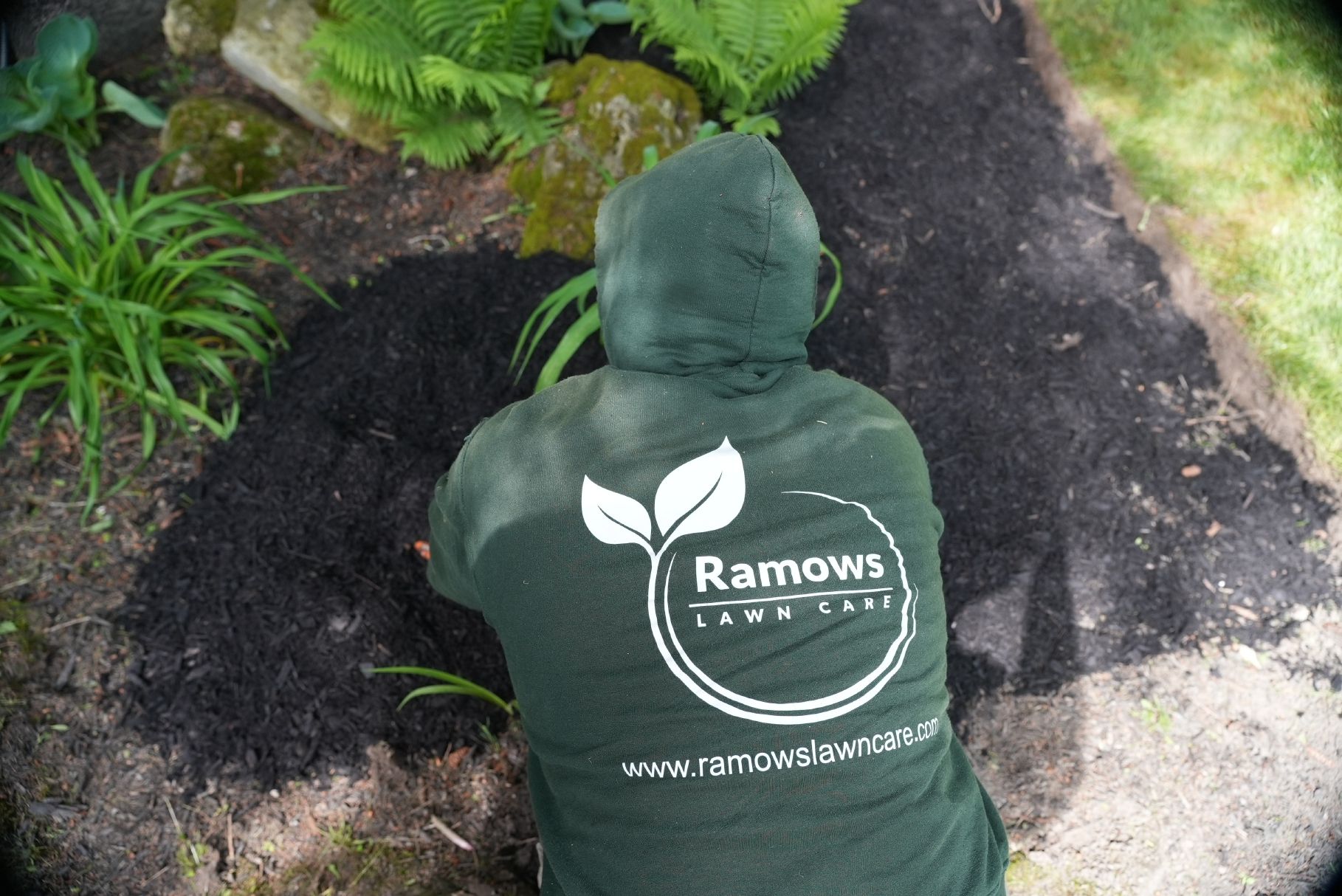 A person in a dark green hooded sweatshirt with the Ramows Lawn Care logo works in a garden bed with black mulch.
