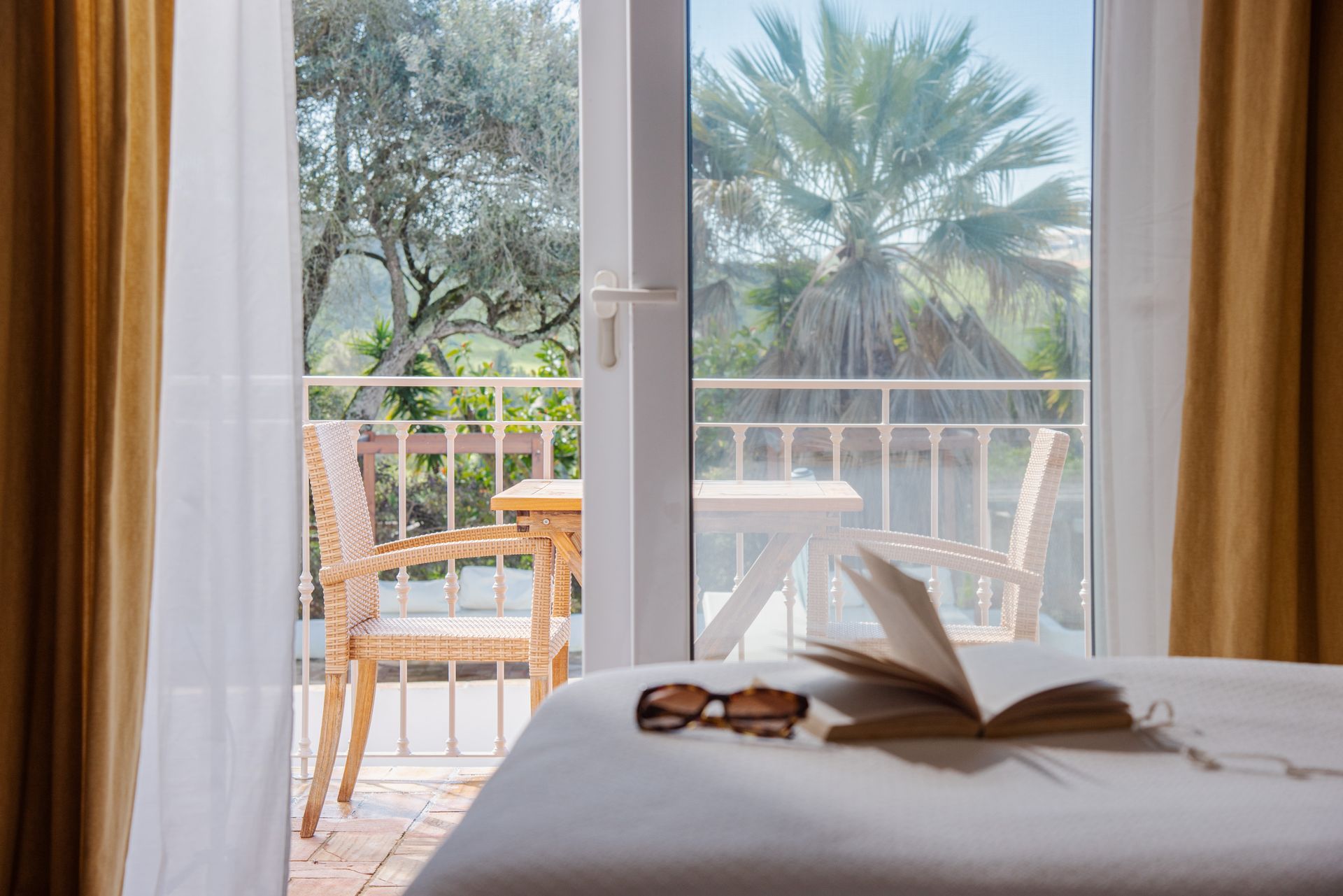 A bed with a book and sunglasses on it next to a balcony.