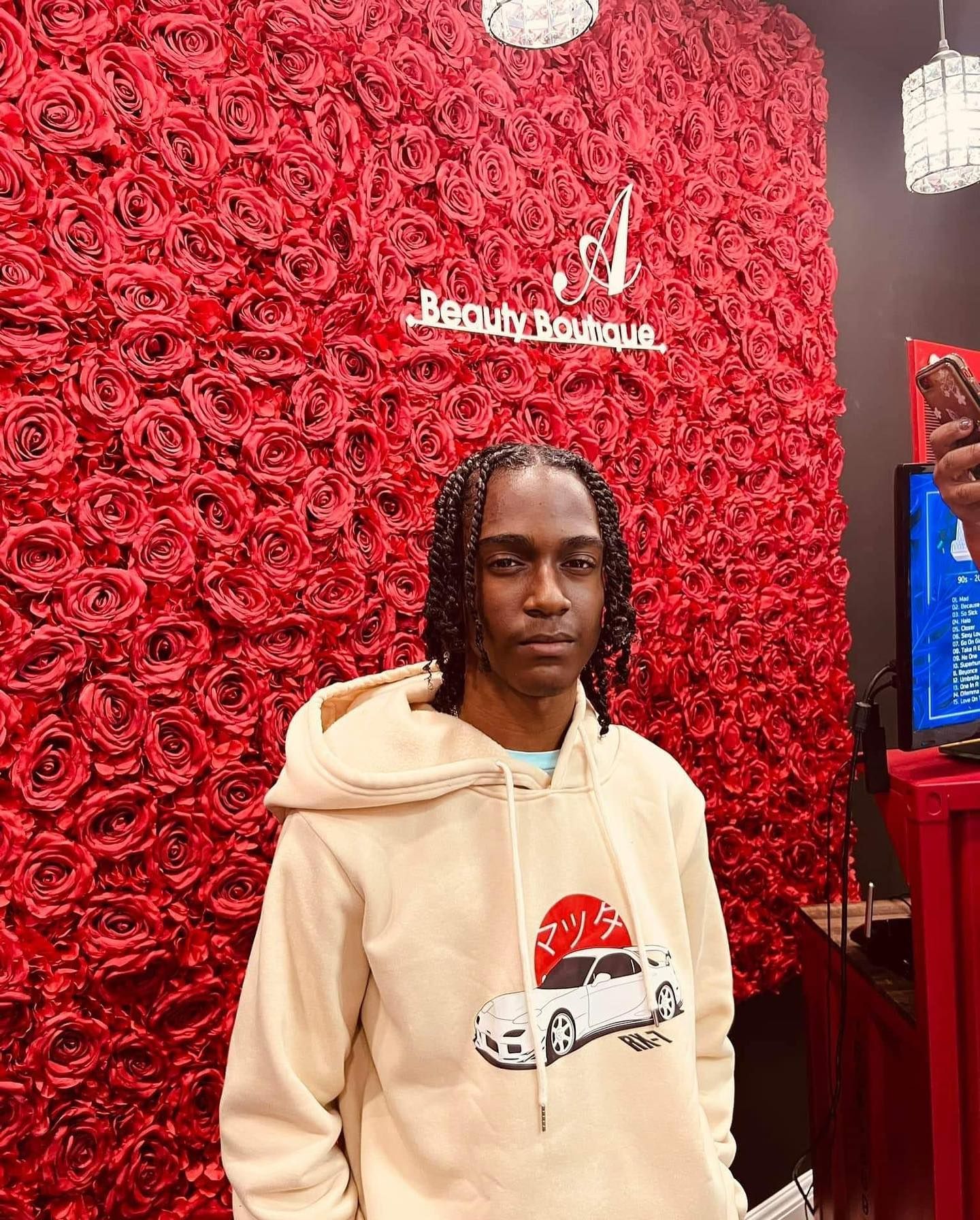 A person with braided hair wears a cream hoodie featuring a graphic of a car against a background of red artificial roses.