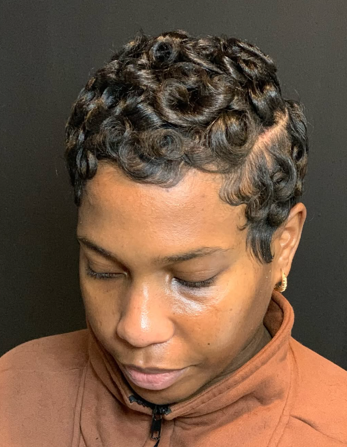 A person with a short, dark hairstyle featuring defined, sculpted curls, wearing a brown zippered jacket.