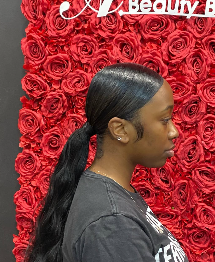 A person with a sleek, low ponytail hairstyle in profile, positioned in front of a decorative red rose wall backdrop.